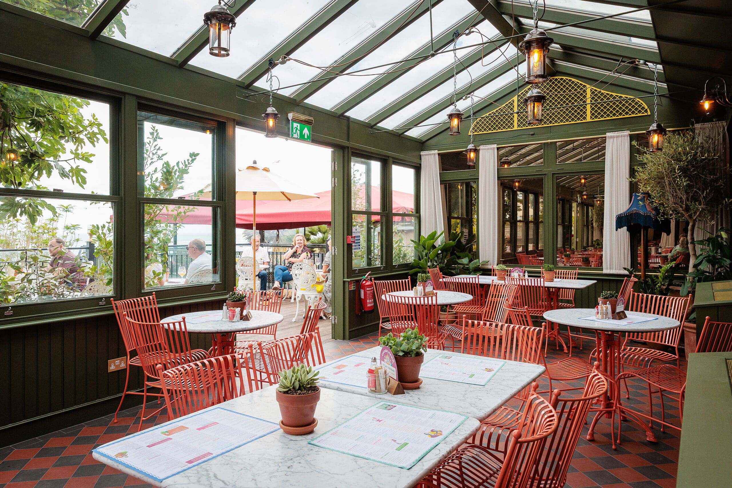 A bright, airy restaurant with a glass ceiling and walls, red metal chairs, and marble tables decorated with small potted plants. A wood-fired forno adds charm as warm lights hang from the ceiling. Outside, people sit under large umbrellas.