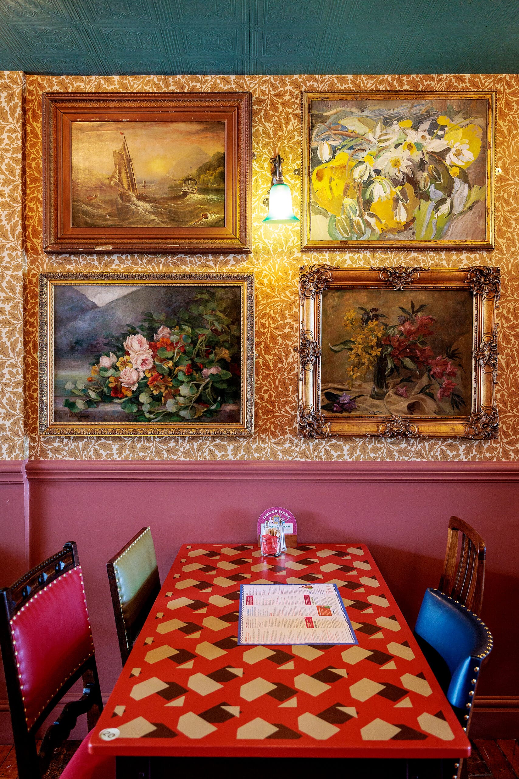 A cozy restaurant table with a red and beige geometric pattern sits near a traditional forno, surrounded by colorful chairs. The wall behind features intricate gold wallpaper and four framed floral and landscape paintings, lit by a single wall lamp.