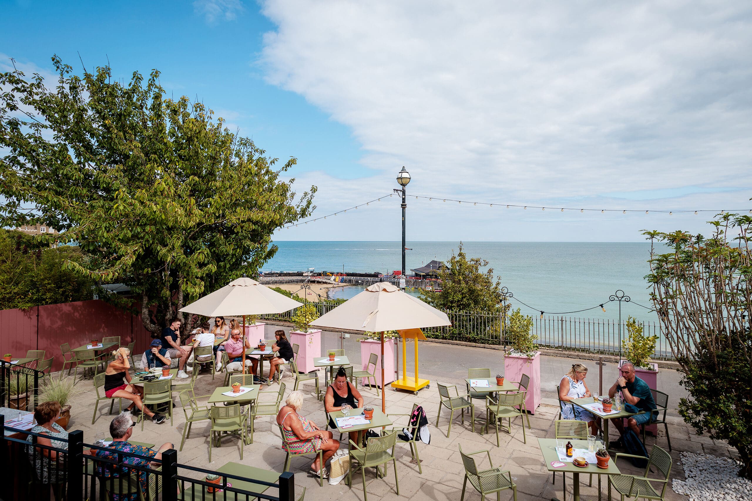 People sit at outdoor tables with umbrellas, savoring food from the forno and drinks on a sunny patio overlooking the sea. Trees, pink walls, and string lights decorate the area under a partly cloudy sky.