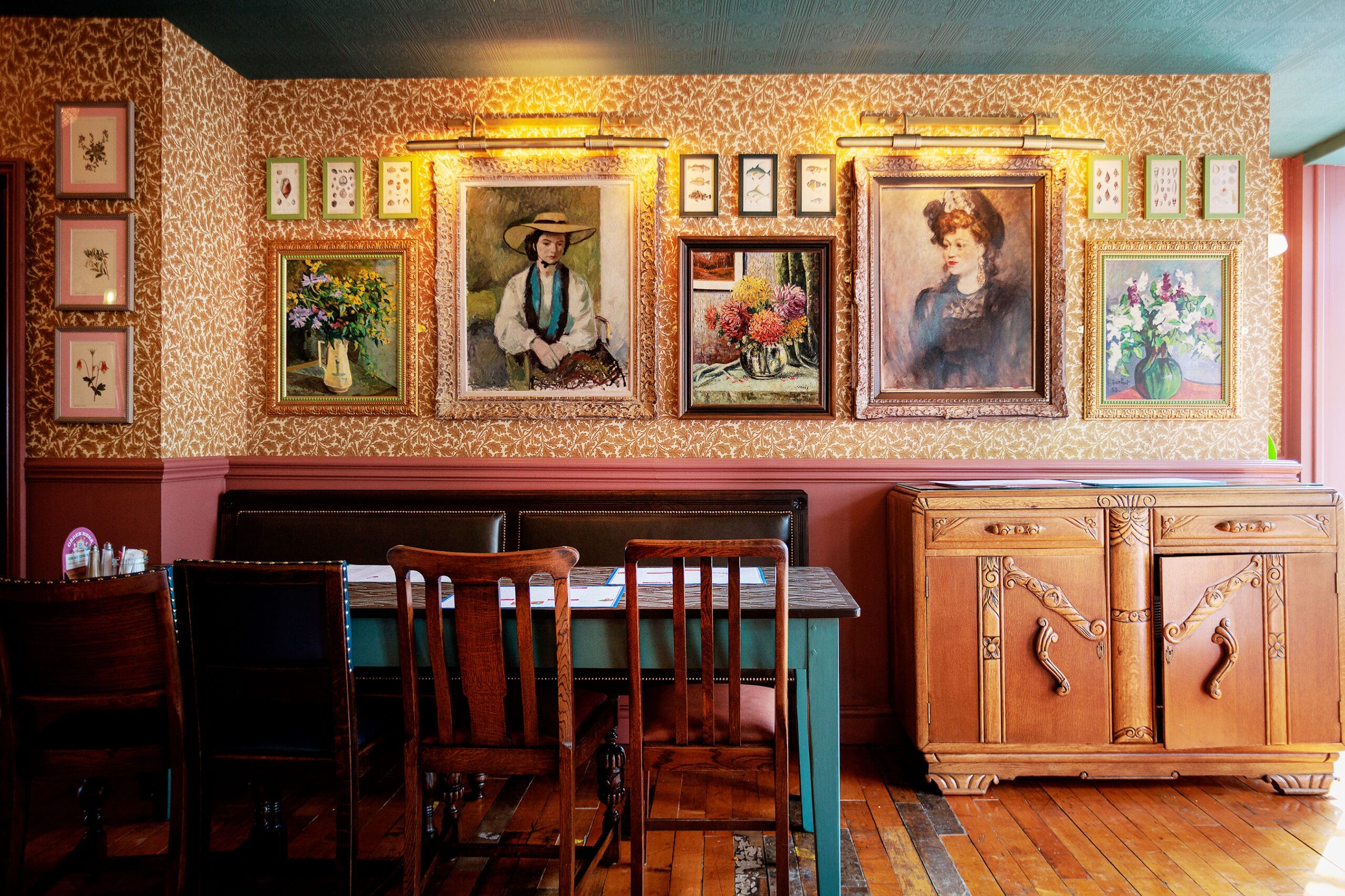 A cozy dining area with vintage wooden chairs and a teal table, floral wallpaper, framed paintings of women and flowers, an ornate wooden sideboard, and a rustic forno adding charm to the inviting space.