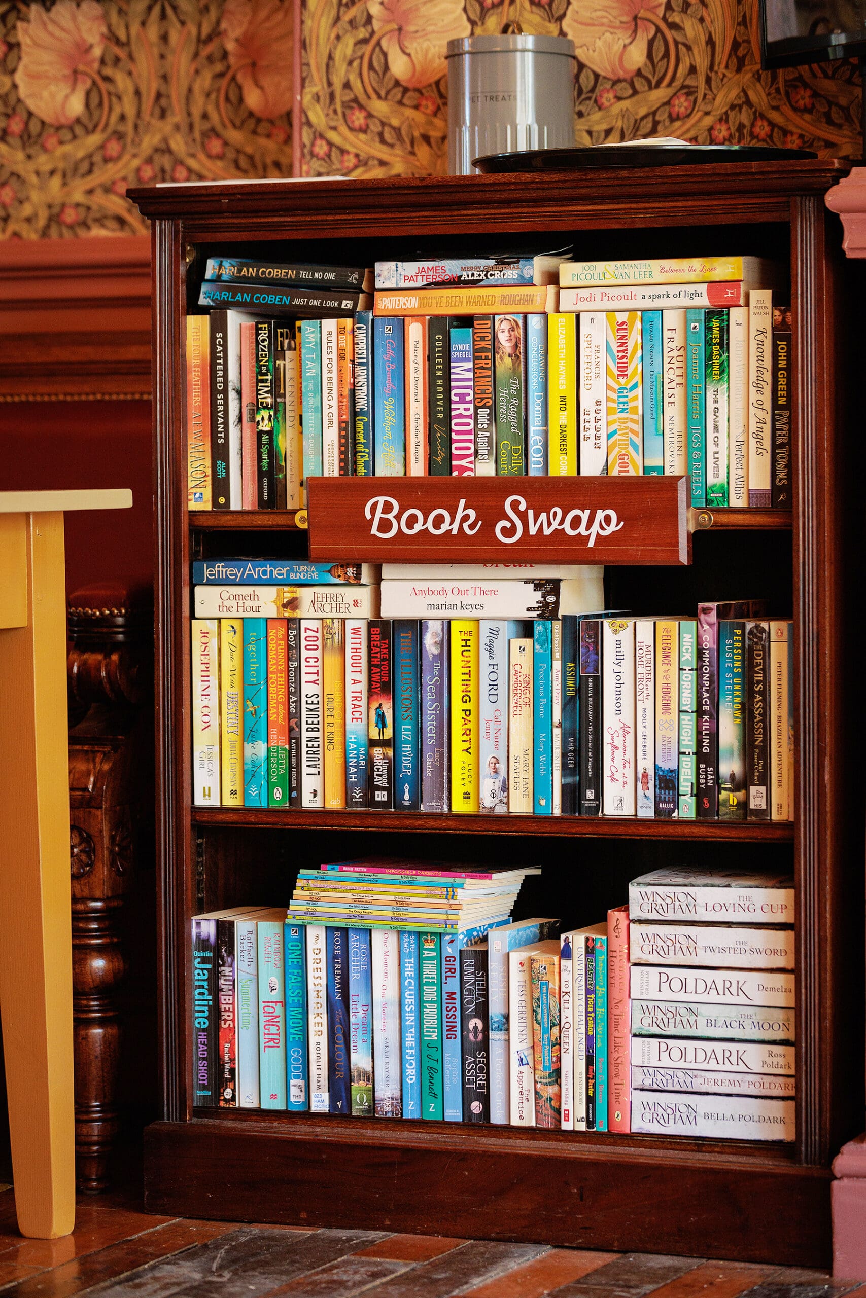 A wooden bookshelf labeled "Book Swap" holds a variety of colorful books, with several stacked horizontally on the bottom shelf. The background features ornate, decorative wallpaper reminiscent of a vintage forno’s cozy charm.