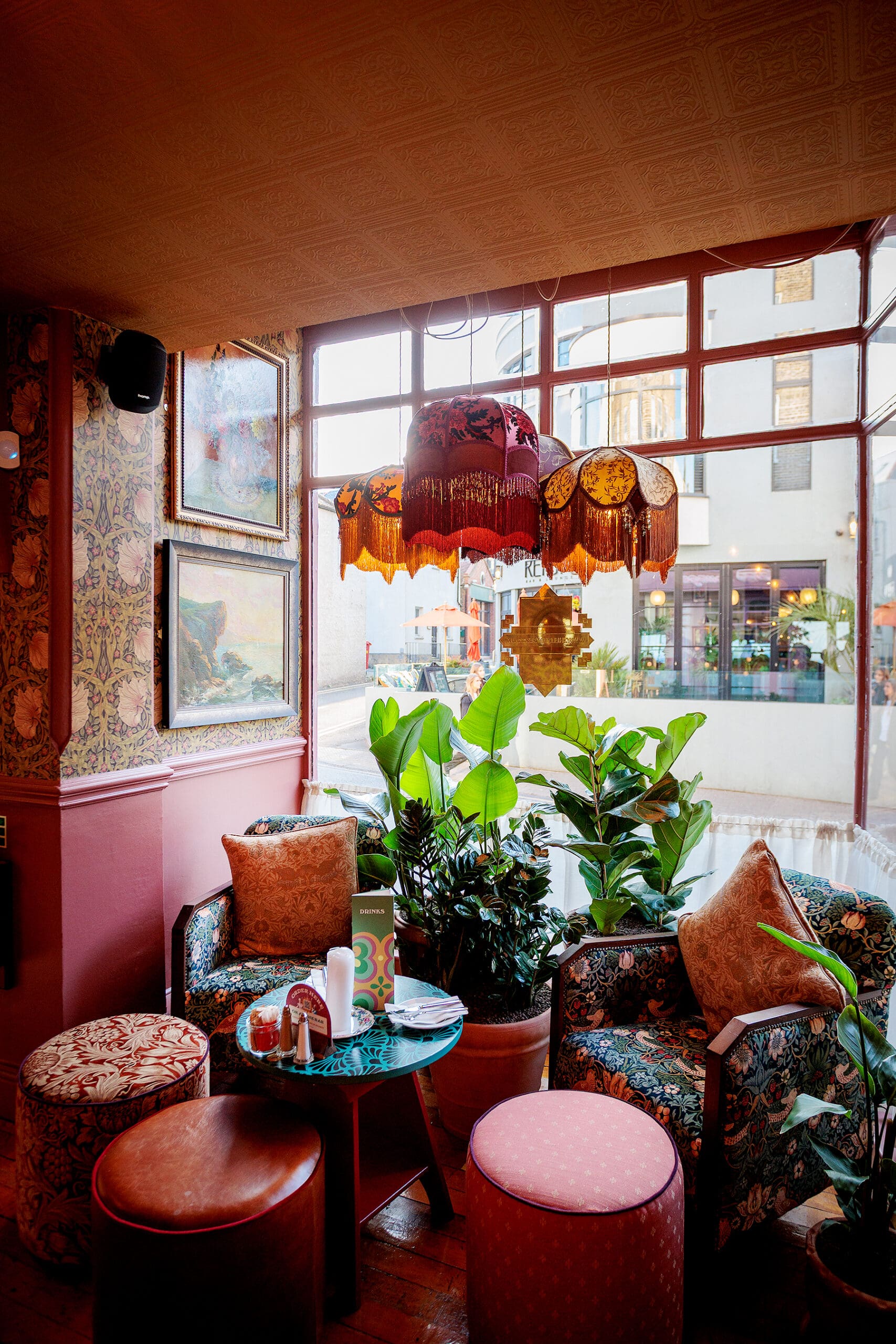 A cozy café corner with patterned armchairs, round ottomans, a small table, and lush green plants features vintage-style lampshades and a charming forno, all set by large windows with city views.