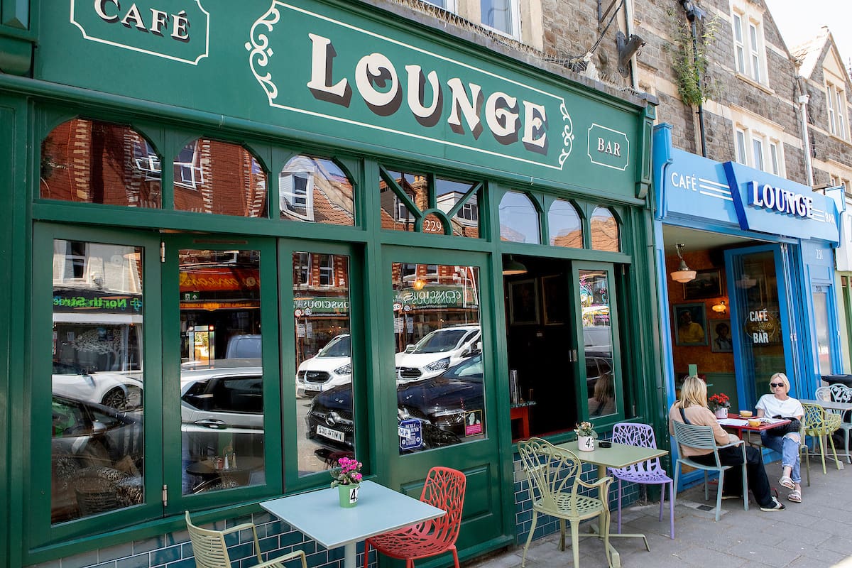 Two people sit at an outdoor table in front of a green café-bar called "Lounge," with colorful chairs and tables on the sidewalk—a welcoming scene that reflects the vibrant spirit shared in our About Us story.