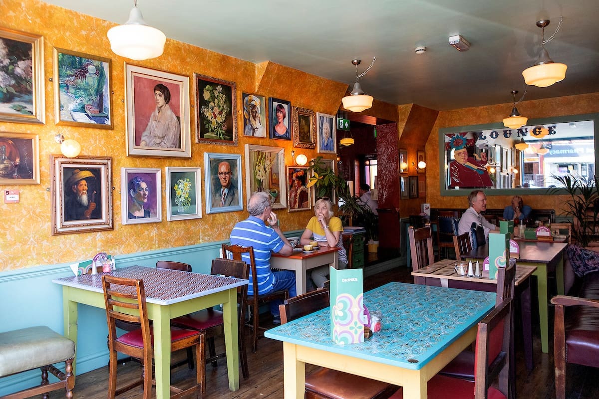 A cozy café, The Lounge, features colorful tables and chairs with people chatting. Its walls are adorned with numerous framed portraits and paintings, creating a warm, eclectic atmosphere bathed in bright natural light from the windows.