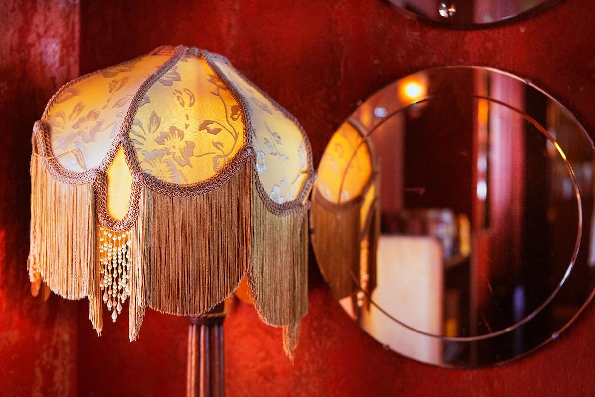 A vintage lamp with a yellow floral lampshade and long fringe stands in The Lounge, set against a red wall with round mirrors reflecting part of the room.