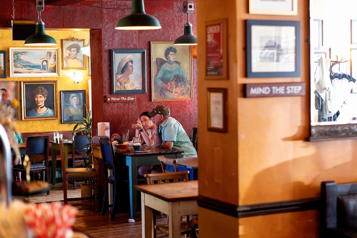 Two people sit and eat at a table in The Lounge, a cozy restaurant decorated with framed portraits on colorful walls. Warm lighting and wooden furniture create a welcoming atmosphere. A "Mind the Step" sign is visible on a pillar.