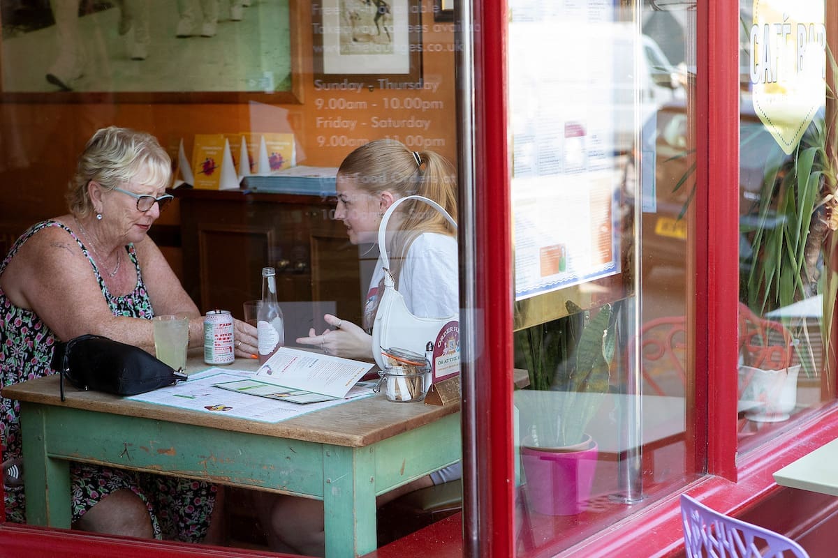 Two women sit at a small table in The Lounge cafe, engaged in conversation. Menus, drinks, and a water bottle are on the table. The scene is viewed through a window, with reflections shimmering on the glass.