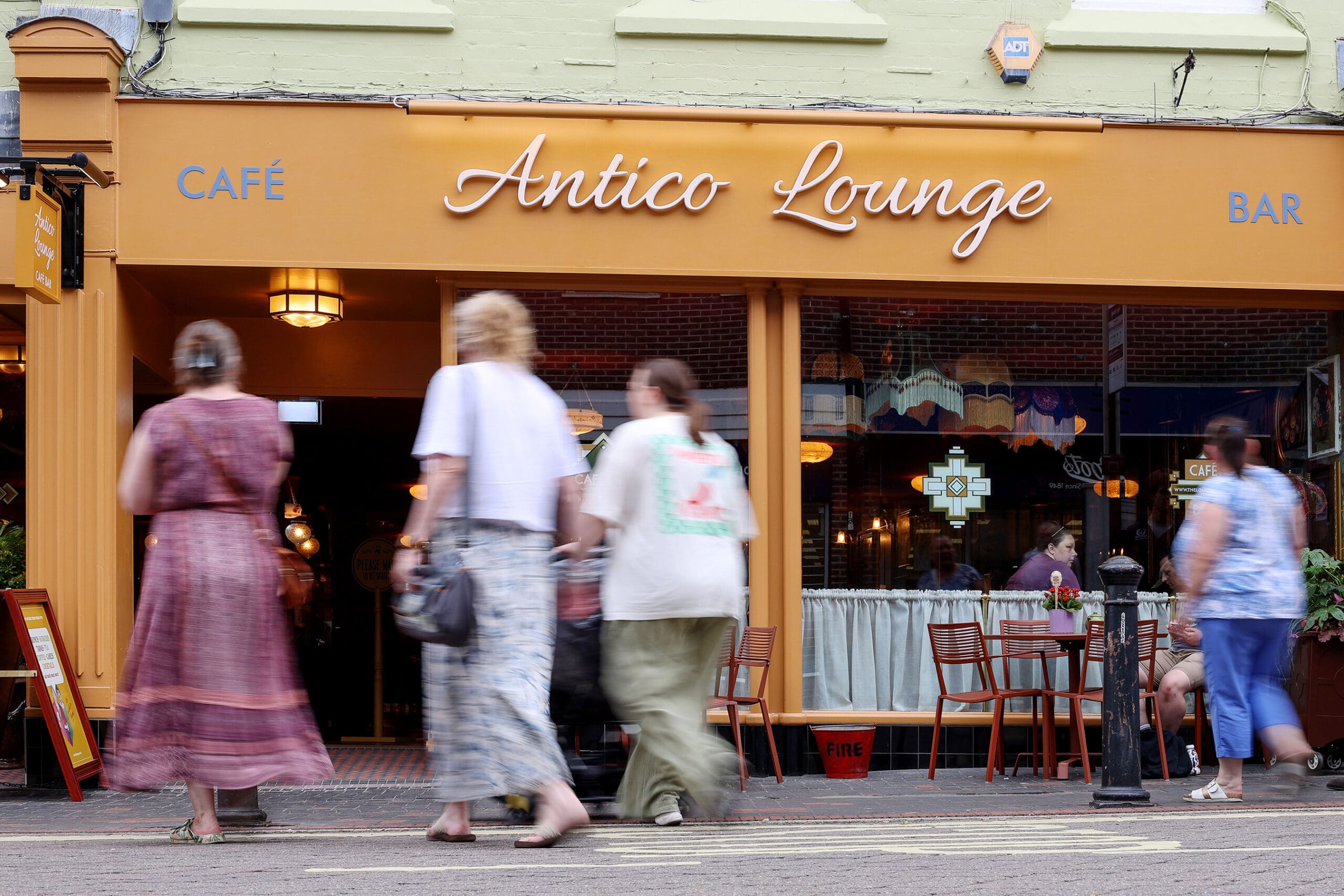 Four people walk past Antico Lounge café and bar with its distinctive orange signage and outdoor seating. Some patrons enjoy the relaxed atmosphere outside, while warm lights illuminate the inviting indoor Antico space.