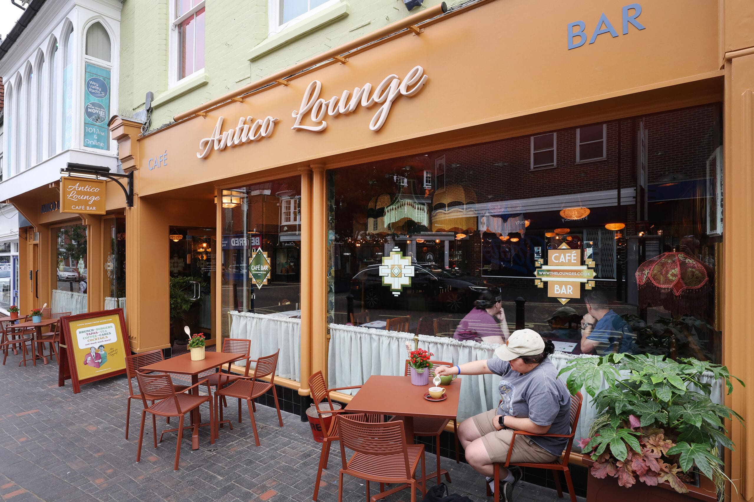 A person sits at an outdoor table with coffee at Antico, enjoying the café’s large windows, orange signage, and potted plants. The street reflects in the glass while more patrons are visible inside this welcoming spot.