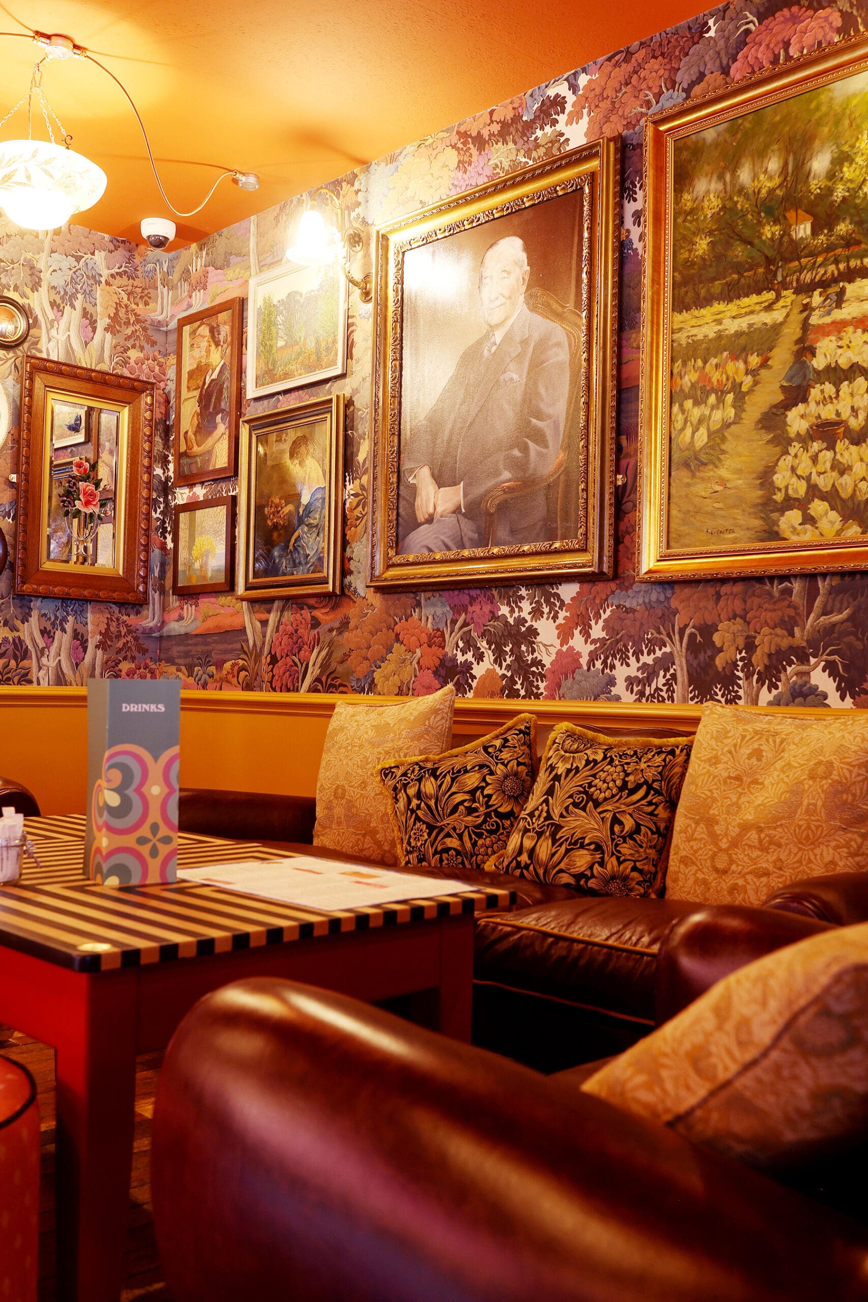 Cozy Antico cafe corner with leather sofas, a wooden table, patterned cushions, and walls decorated with colorful floral wallpaper and framed paintings, including a large portrait of a man. Warm lighting creates an inviting atmosphere.