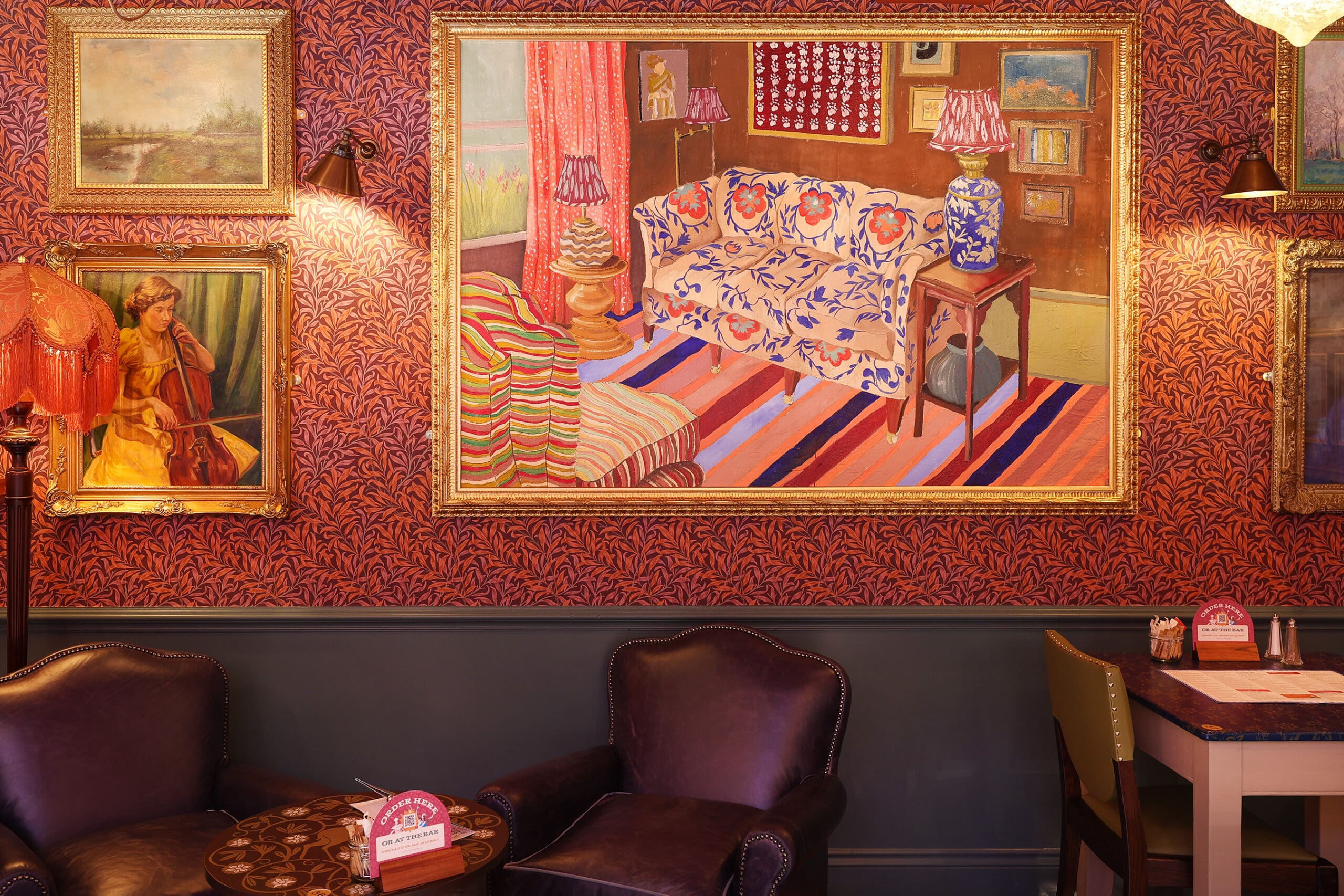 A cozy Antico room with ornate red wallpaper, leather armchairs, a small table, and a dining table. Large framed paintings, including one of a colorful living room, adorn the walls. Warm ambient lighting creates an inviting atmosphere.