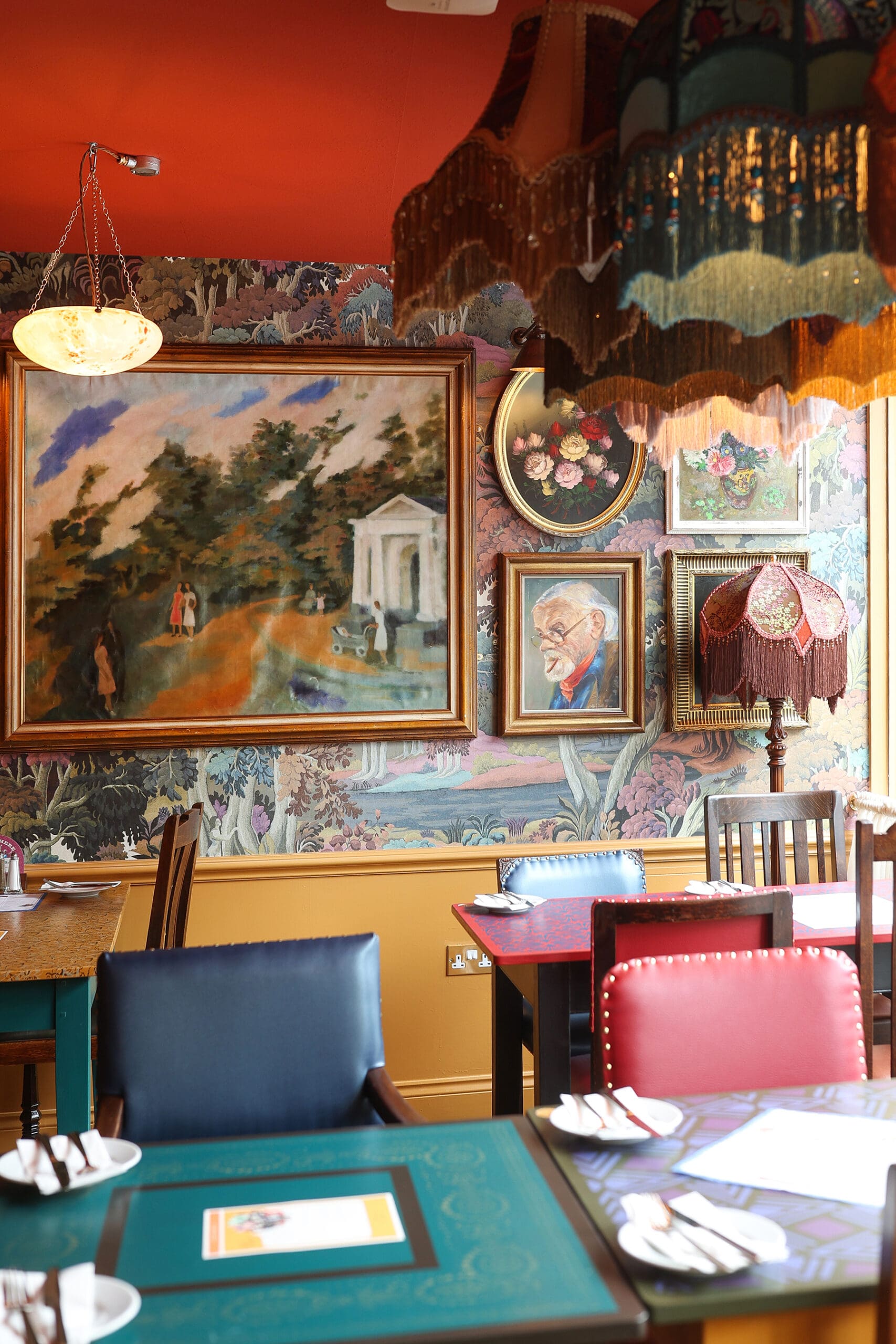 A cozy, colorful restaurant interior with eclectic Antico decor, mismatched chairs, vibrant tables, and various framed paintings and portraits on patterned wallpapered walls under ornate lampshades.