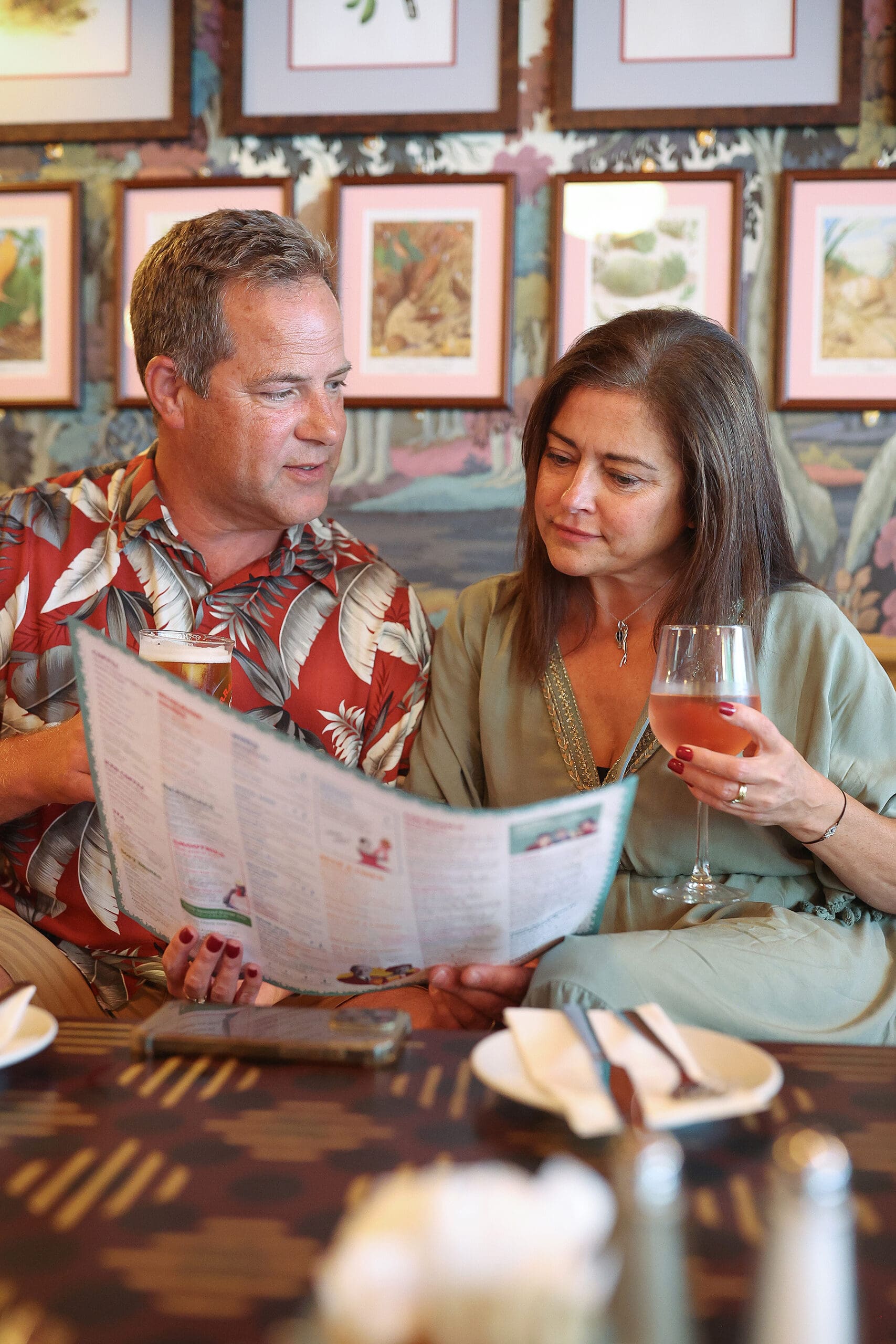 A man and woman sit closely together at Antico, holding drinks and looking at a menu. Framed art decorates the wall behind them, and a table with plates, napkins, and cutlery is in the foreground.
