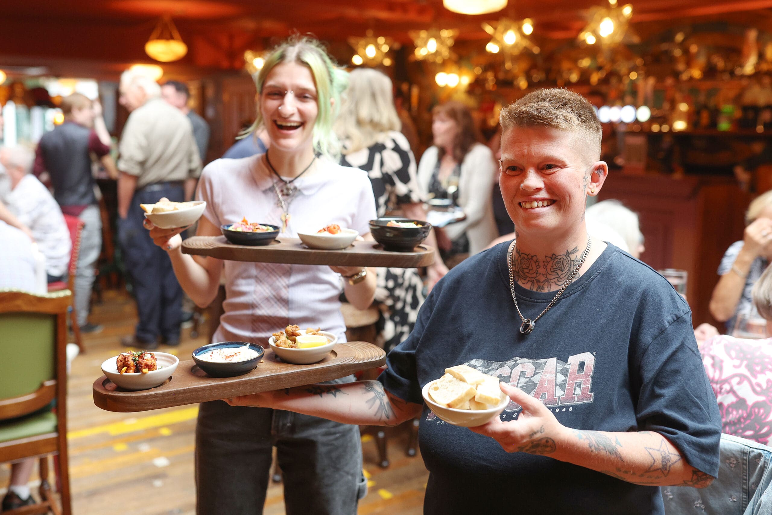 Two smiling people in the lively Antico restaurant hold wooden trays with small bowls of food. The bustling atmosphere is filled with seated guests chatting and enjoying their meals in the background.