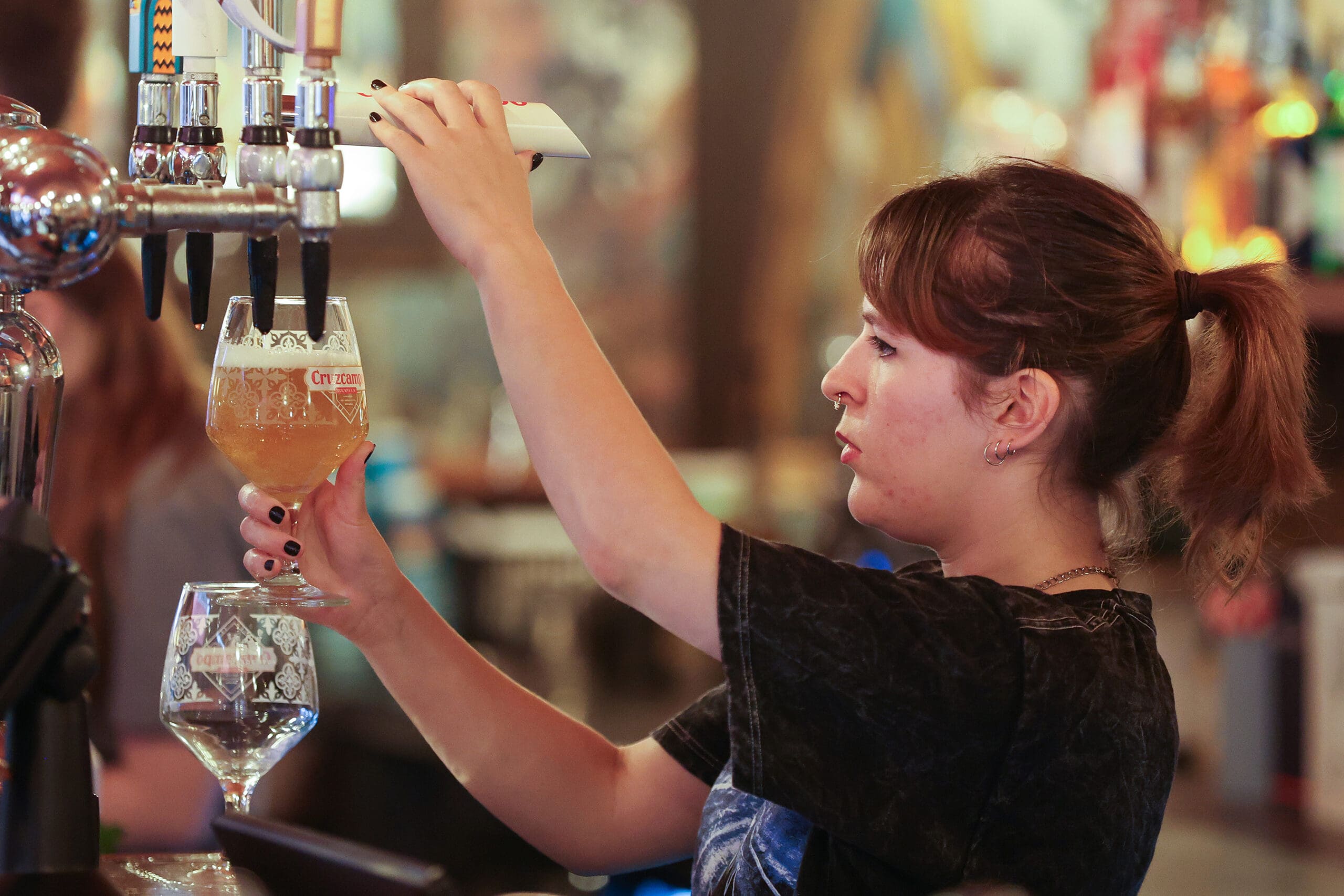 A bartender at Antico pours a glass of beer from a tap, focusing on filling the glass, with another empty glass nearby on the bar counter. The background is softly blurred.