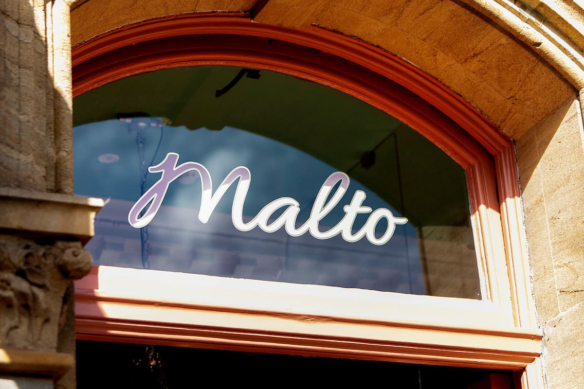 A glass window with the word "Malto" elegantly scripted in white, set in a stone building with a reddish-brown arched frame.