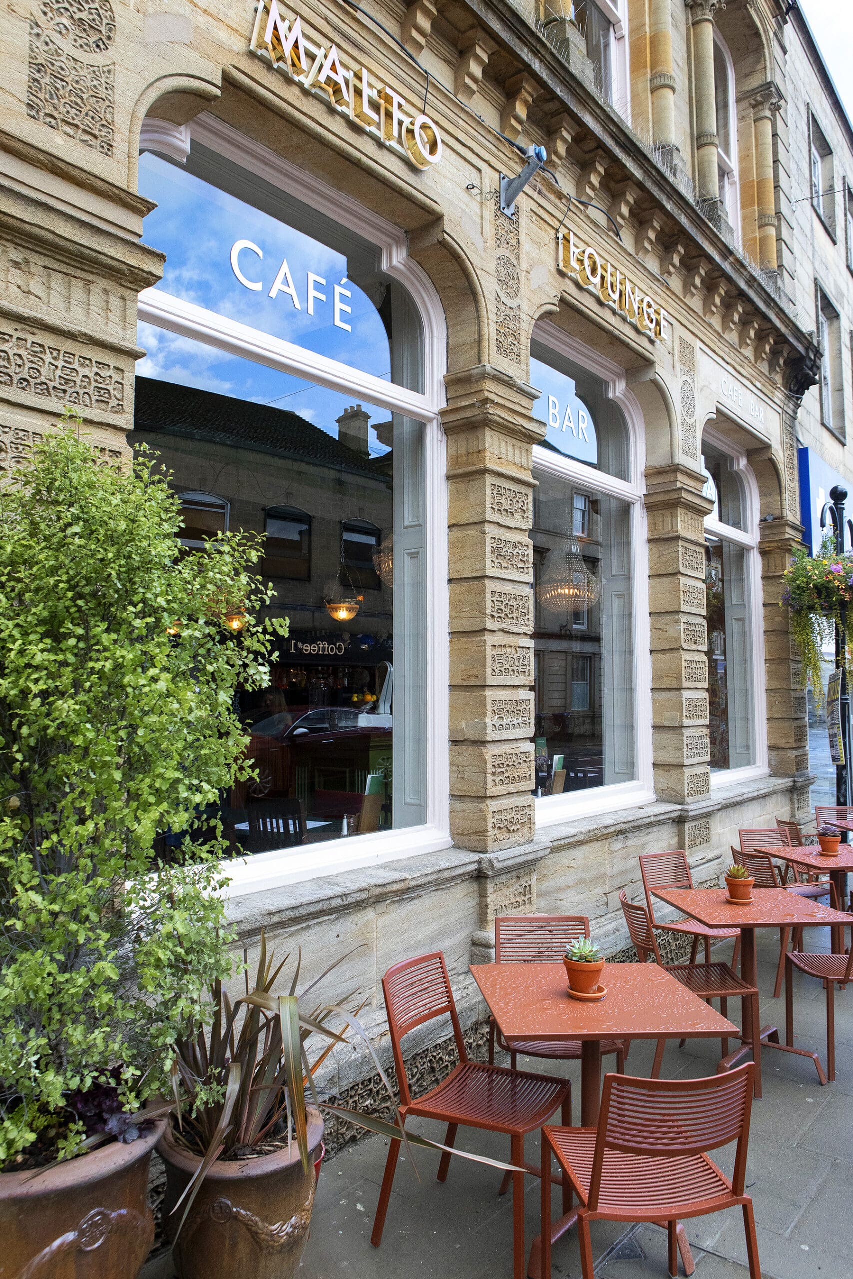 Outdoor seating with red tables and chairs in front of Malto café and lounge. Large windows reveal indoor lights and greenery, while potted plants decorate the sidewalk for a welcoming atmosphere.