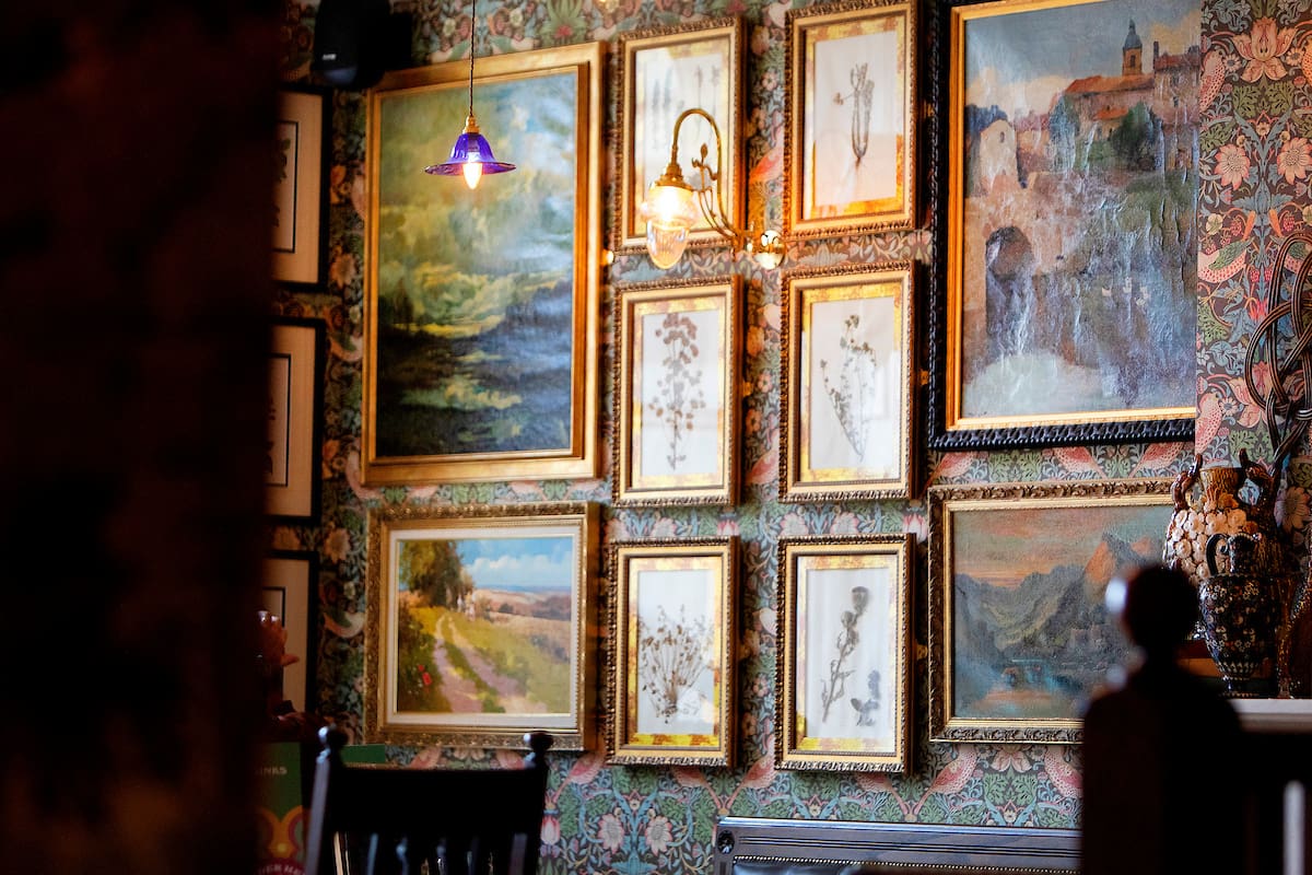 A warmly lit room with Malto floral-patterned wallpaper and numerous framed paintings and botanical prints hanging on the wall, featuring landscapes and plants. A wooden chair is visible in the foreground.