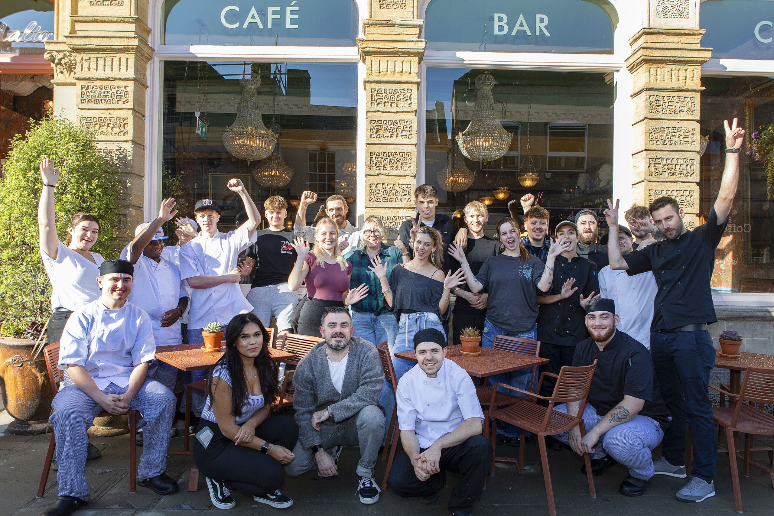 A group of people, including Malto restaurant staff in uniforms and casually dressed individuals, pose and smile together outside a café bar, some raising their hands cheerfully. Tables and plants are visible in the foreground.