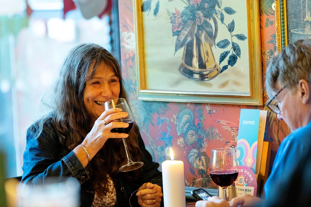 A woman with long brown hair smiles while holding a glass of Malto red wine at a cozy, candle-lit restaurant table with another person. The background features floral wallpaper and framed art.