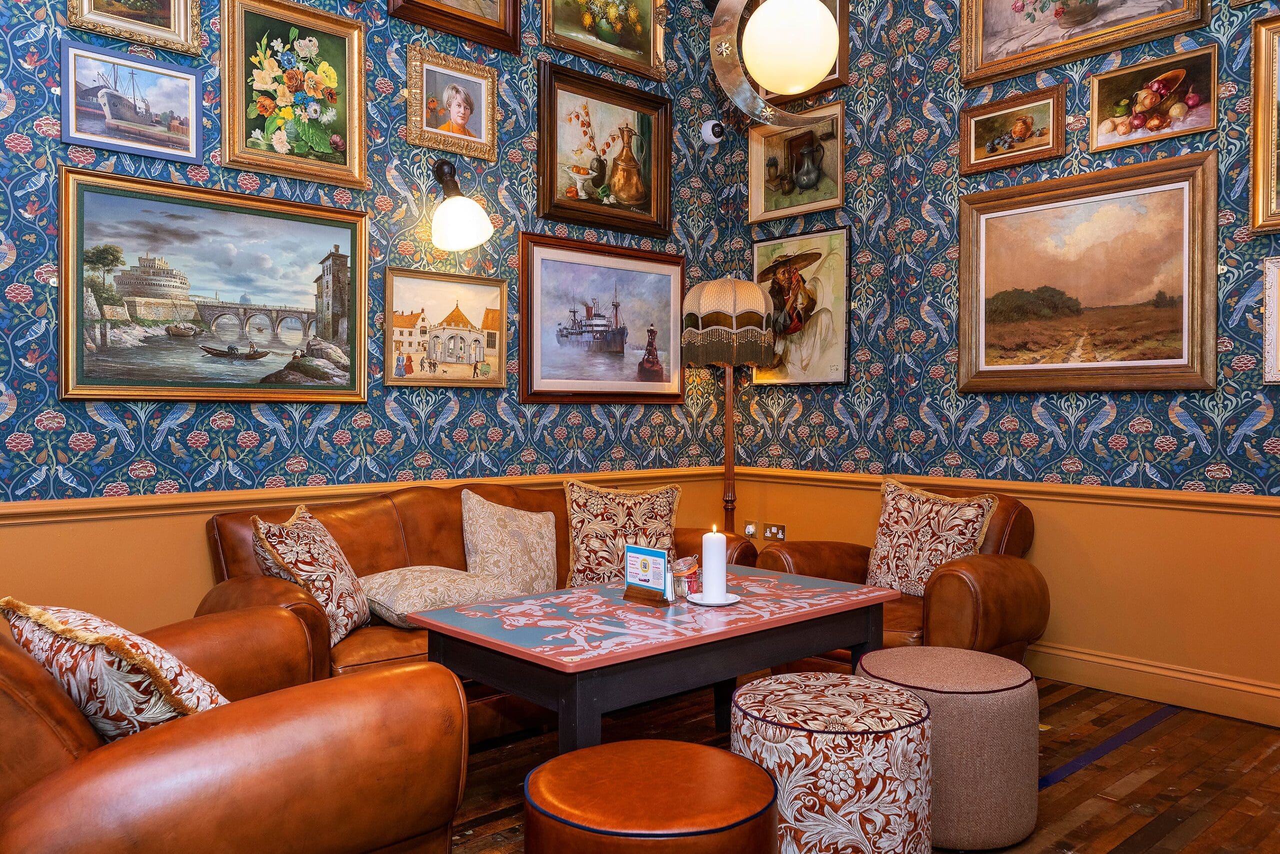 Cozy café corner with brown leather chairs, patterned cushions, floral wallpaper, and framed paintings. A decorated wooden table and two round ottomans rest on a wooden floor, creating a warm, inviting piccolo lounge ambiance.