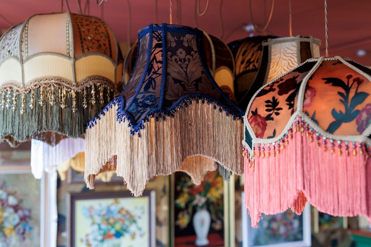 A group of vintage lampshades with colorful fringe and decorative patterns hang from the ceiling, adding a sarto-inspired touch, with framed floral paintings blurred in the background.