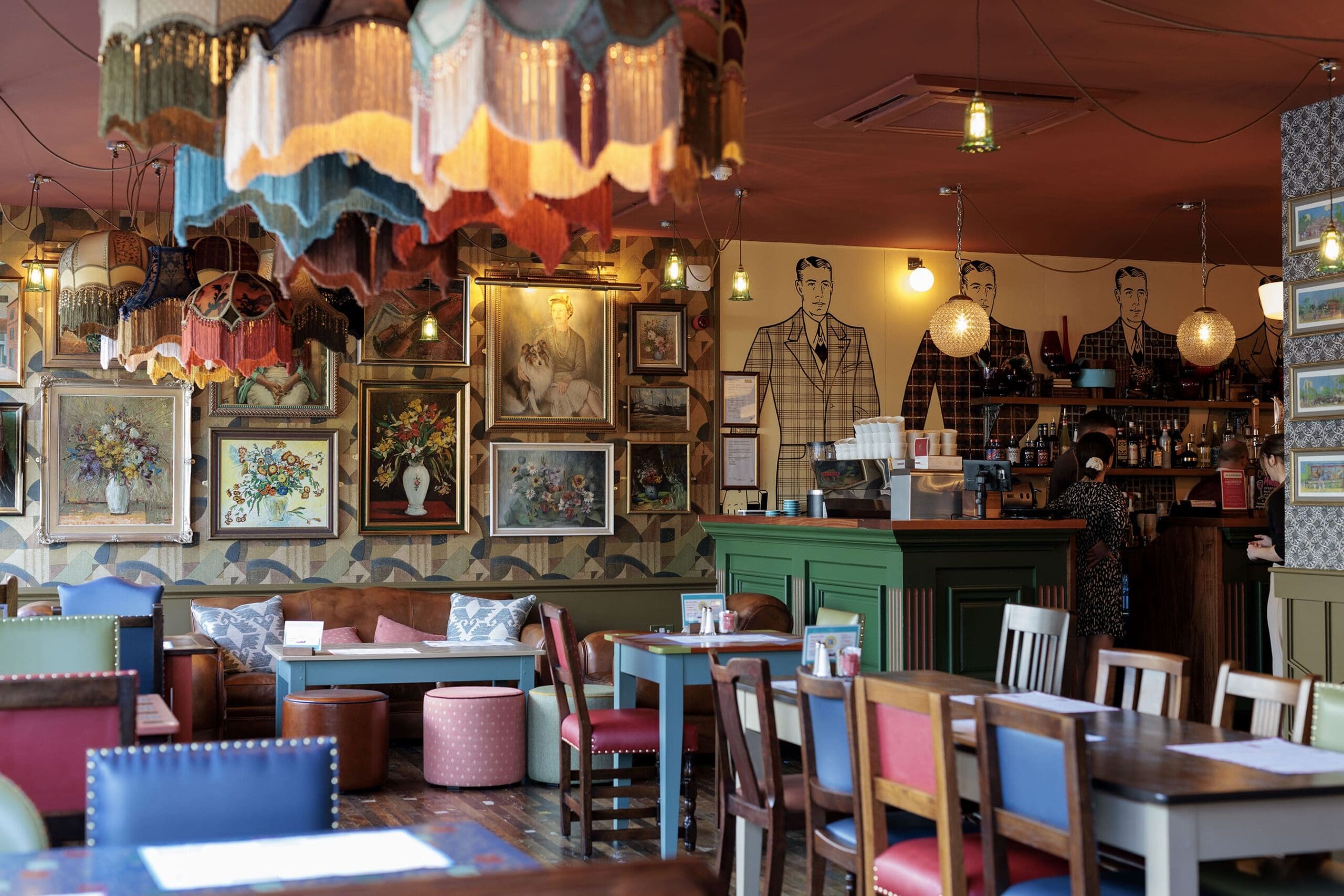 Eclectic restaurant interior with colorful chairs, vintage lamps, and framed artwork on the walls. The bar area features a green counter and sarto-inspired wall art of men in plaid suits behind it. The space feels cozy and vibrant.