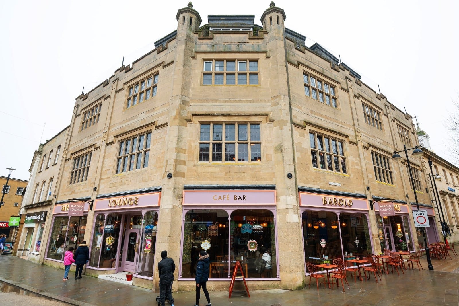 A large, beige stone corner building with tall windows houses Barolo Lounge café bar. The distinctive Barolo pink signage and outdoor seating are visible; people walk and sit outside on a wet, overcast day.