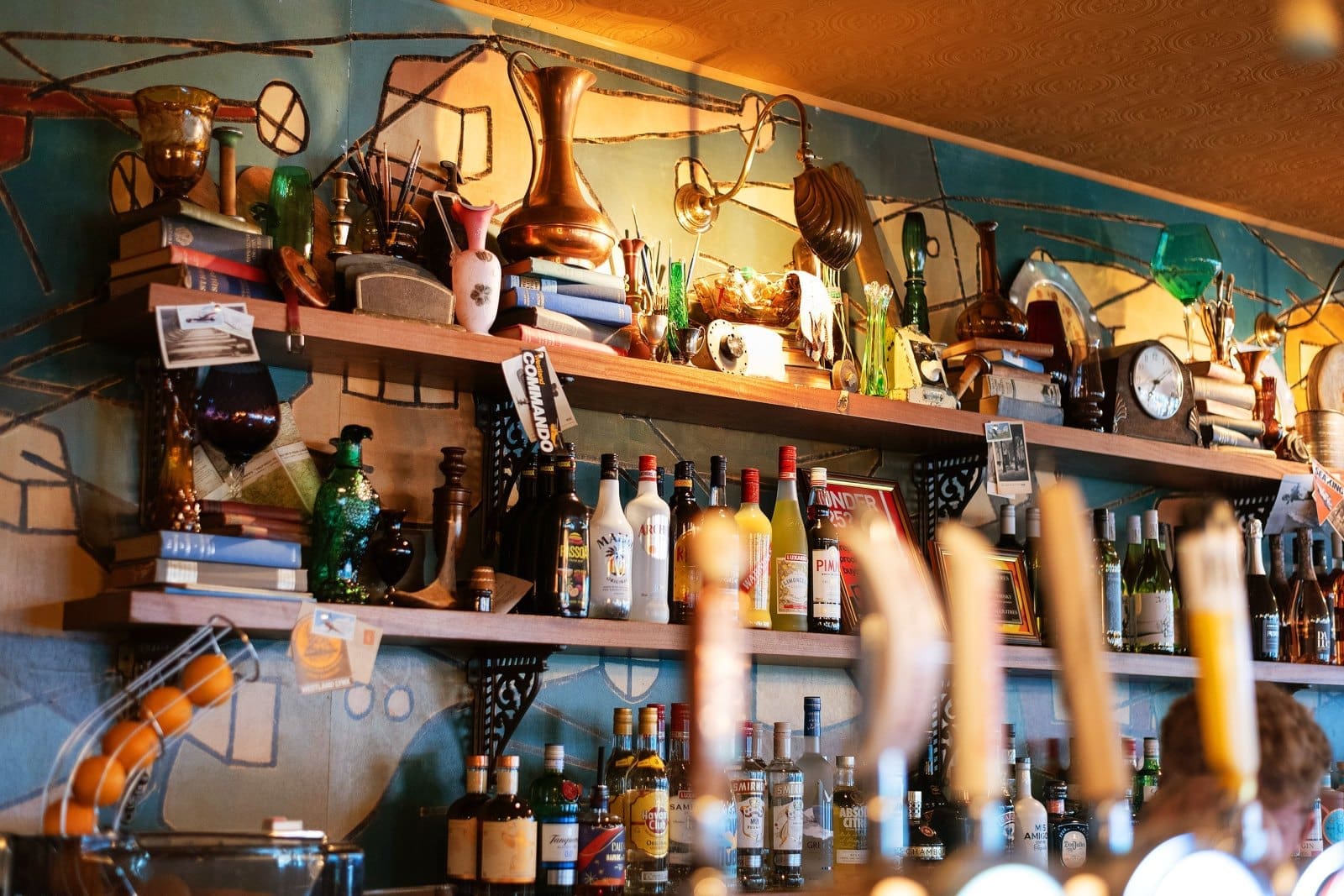 Colorful, cluttered bar shelves filled with Barolo bottles, books, glassware, vintage objects, and quirky decor against a bright mural background; liquor bottles and taps are visible in the foreground.