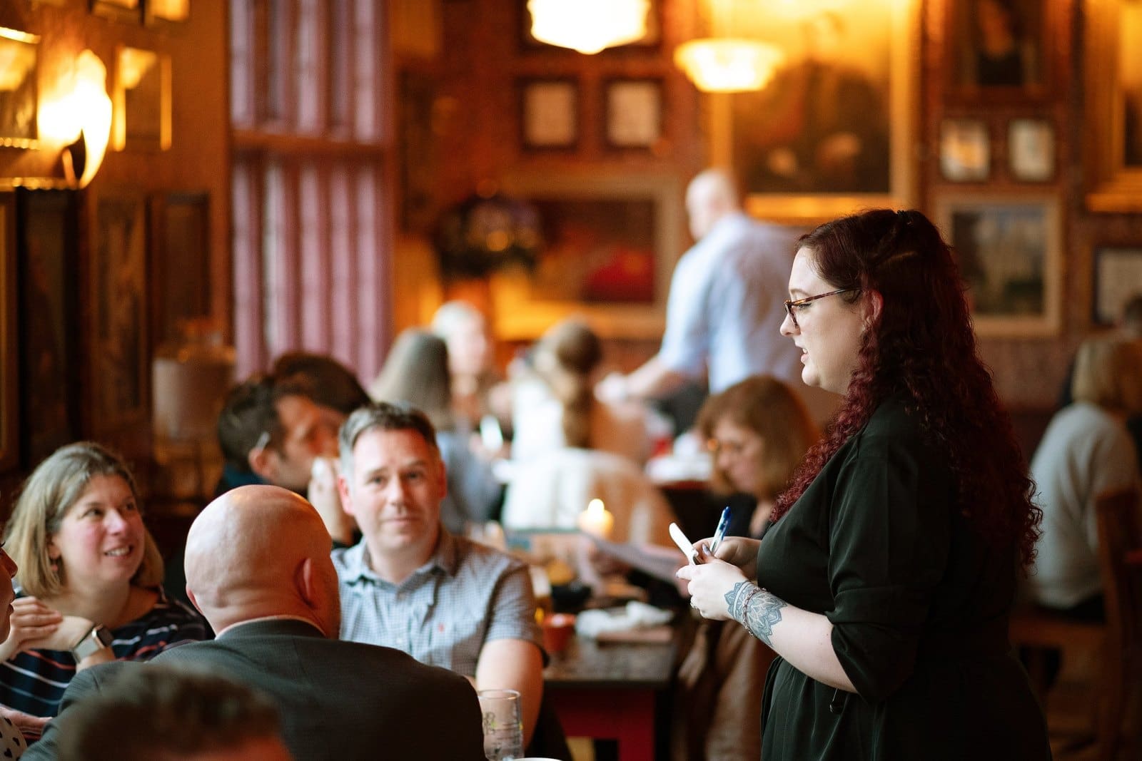 A waitress with long curly hair takes an order from a group of people seated at a restaurant table, as other diners chat in the warmly lit, busy background and glasses of Barolo await on nearby tables.
