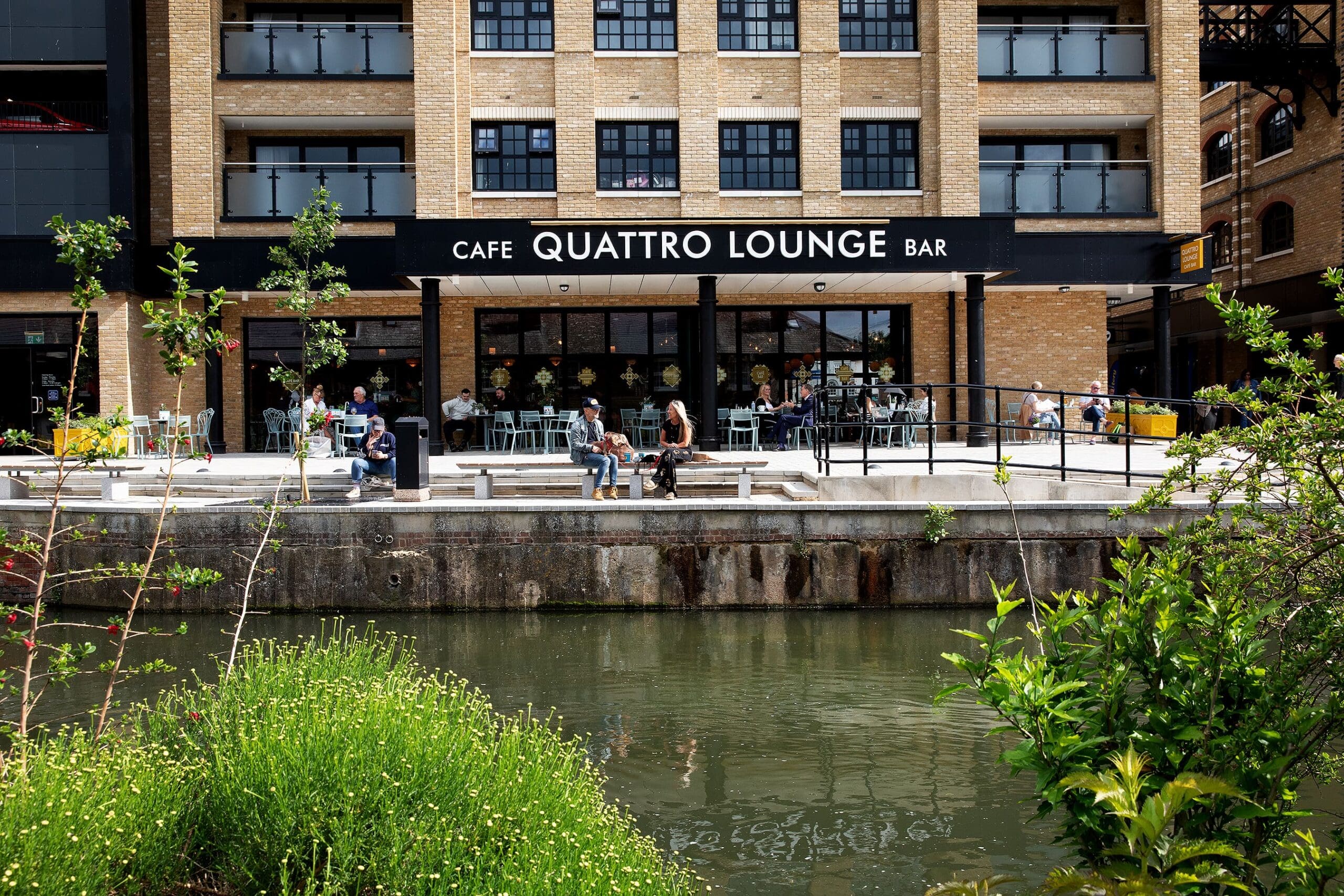 People sit and relax outside Quattro Lounge Bar, a modern building with large windows, outdoor seating, and greenery, next to a calm canal reflecting the vibrant Quattro scene.