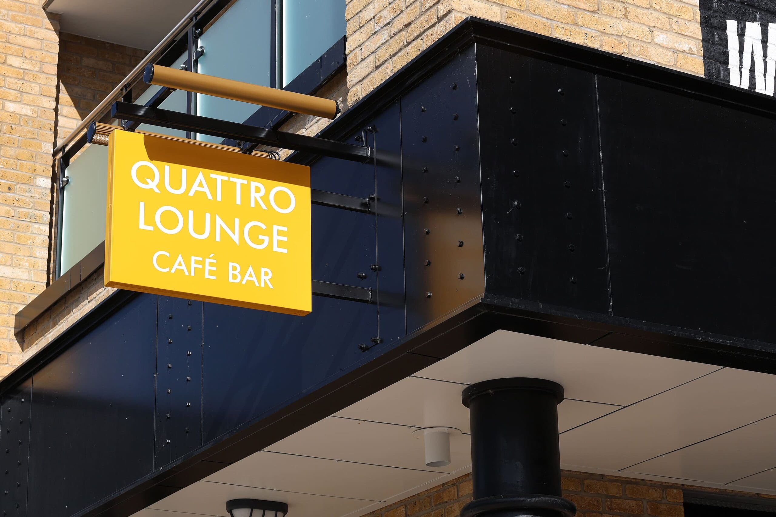 Yellow "Quattro Lounge Café Bar" sign mounted on the exterior of a modern brick and black building, photographed from below during daylight. The bold Quattro branding stands out against the contemporary architecture.