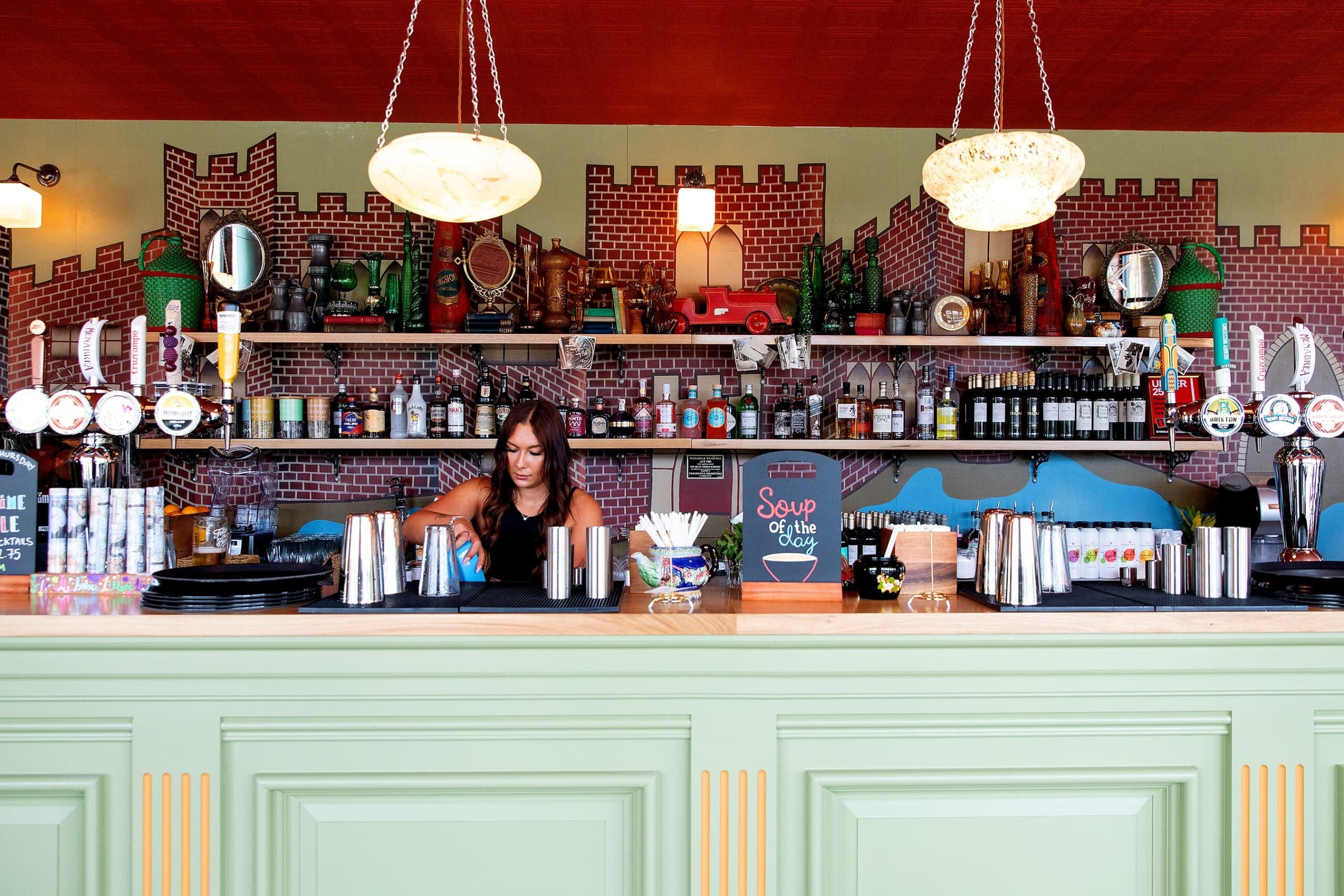 A bartender prepares drinks behind a pastel green Quattro bar counter, with a display of bottles, bar tools, and quirky decorations, including a toy castle and a chalkboard reading “Soup of the Day.”.