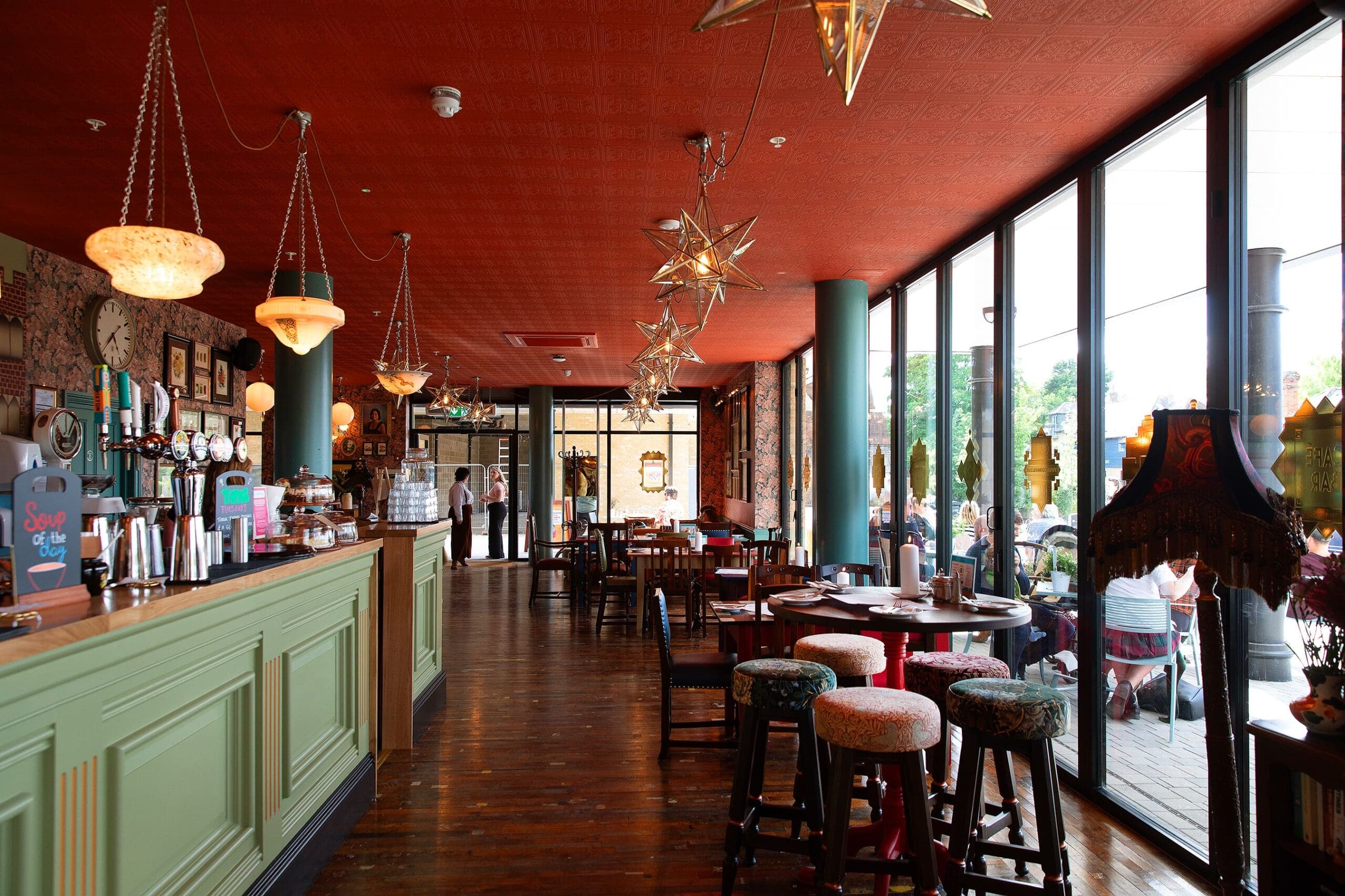 A cozy café with a green wooden bar, round tables, Quattro upholstered stools, star-shaped pendant lights, and large windows letting in natural light. People are seated outside, and the interior has vintage décor.
