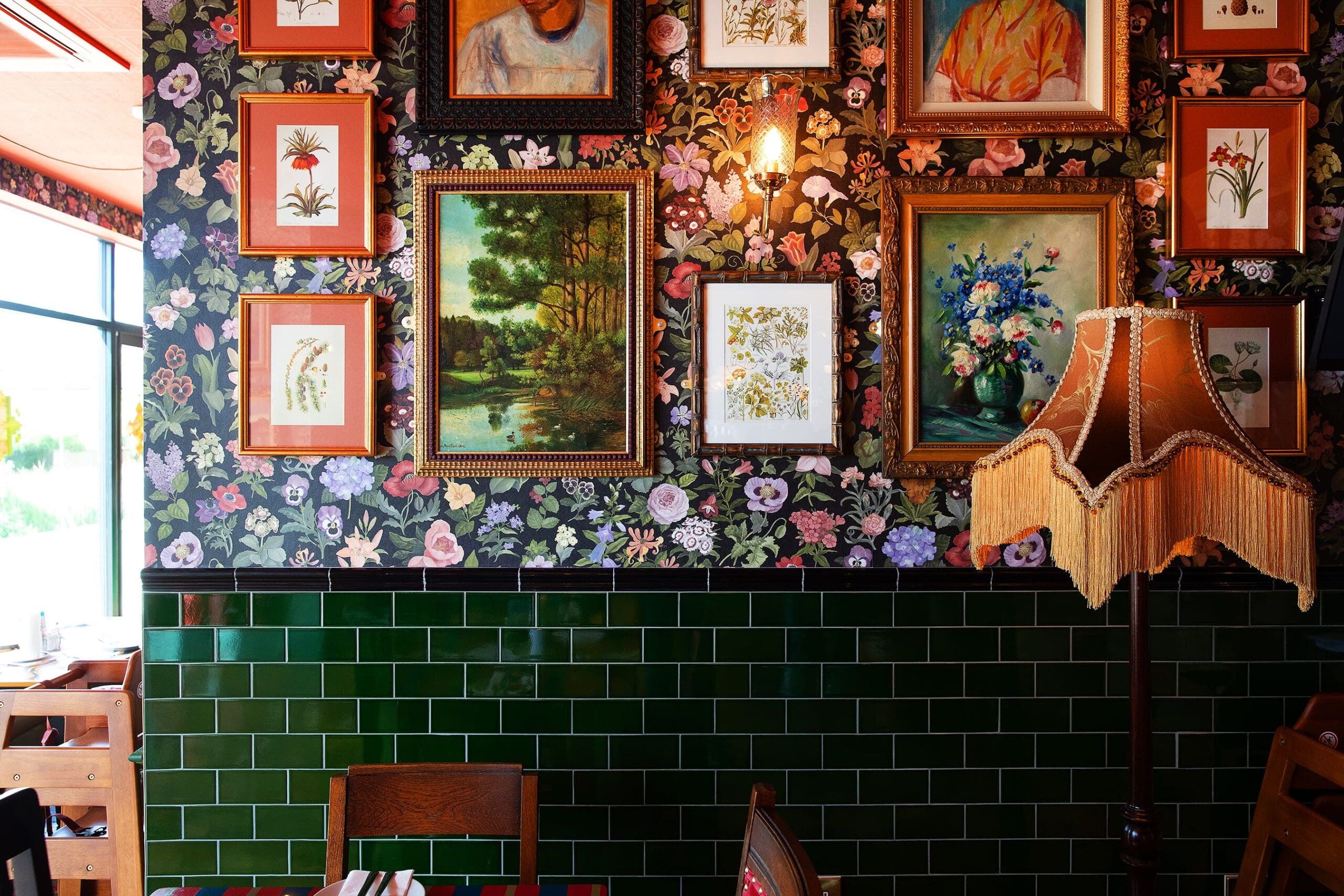 A cozy Quattro restaurant interior with floral wallpaper, green tile walls, framed art and botanical prints, and a vintage lampshade with fringe beside wooden tables and chairs.