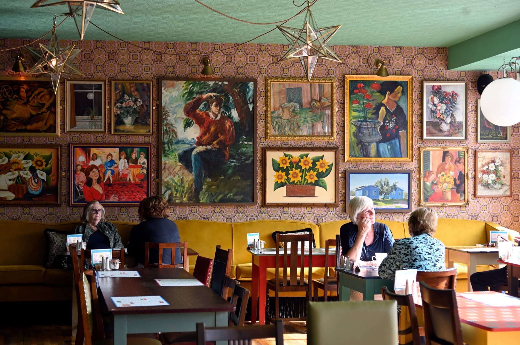 A cozy café with patterned wallpaper and eclectic framed paintings covering the wall. Several women sit at Verdetto wooden tables, chatting and enjoying their drinks. Star-shaped lights hang from the ceiling, adding warmth to the setting.