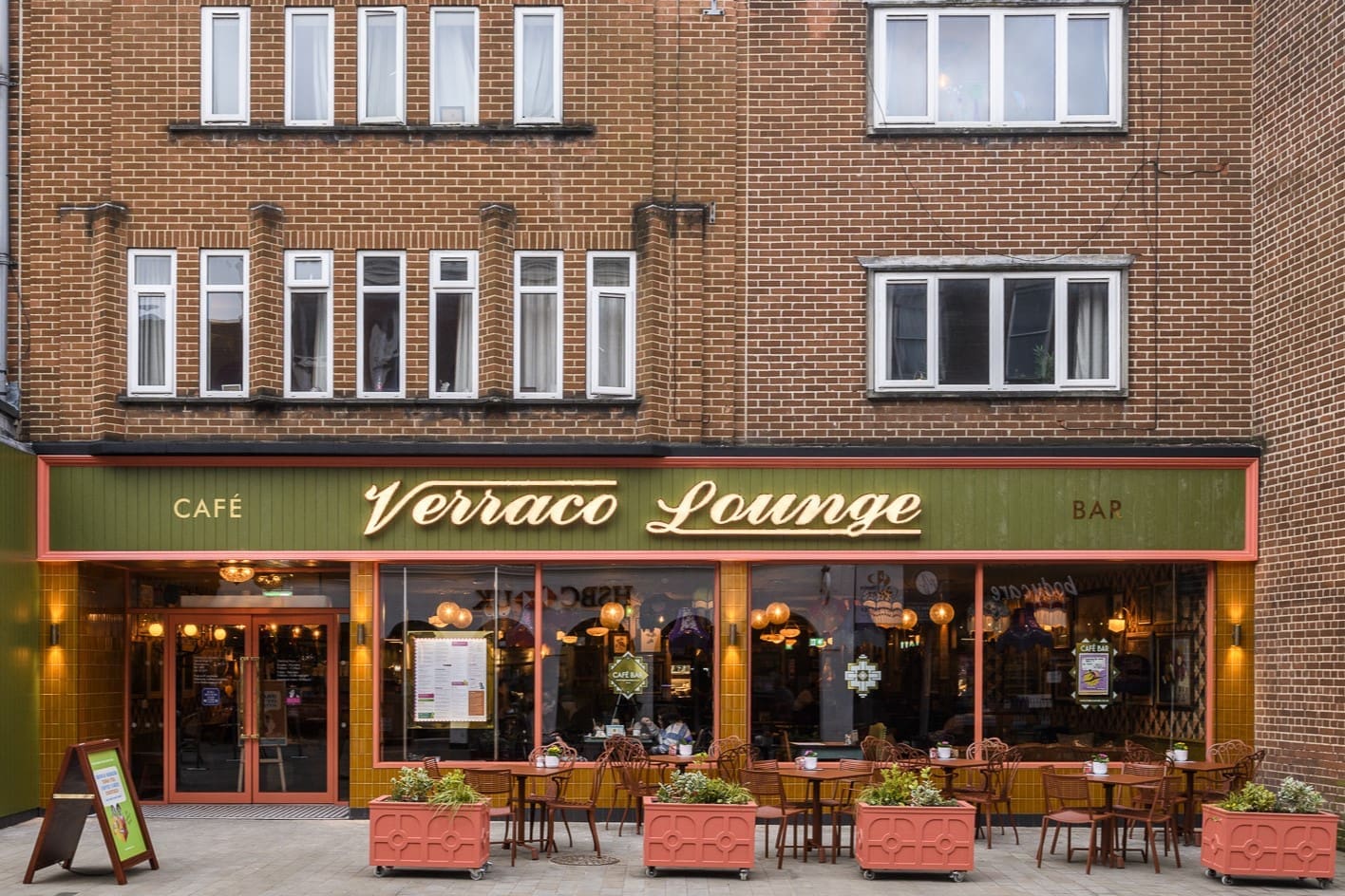 A street view of Verraco Lounge café and bar, featuring large windows, stylish outdoor seating with tables and chairs, and pink planters with green plants decorating the entrance to the verraco-themed brown brick building.