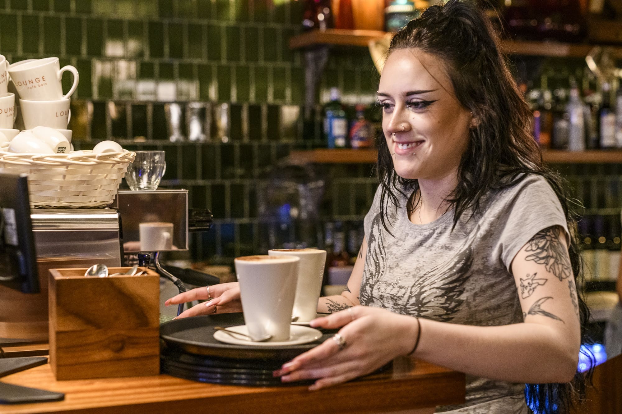 A smiling, tattooed barista with dark hair places two cups of verraco coffee on a tray at a café counter, with mugs and shelves in the background.
