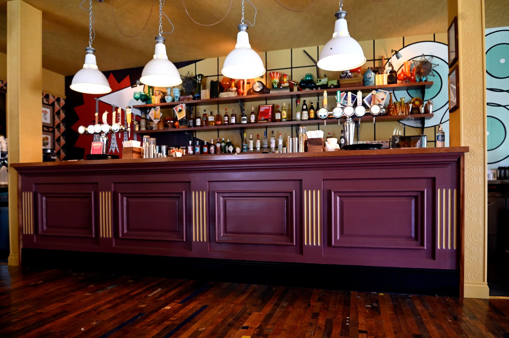 A stylish bar, Viento Lounge, features a dark maroon wooden counter, white pendant lights above, and shelves stocked with bottles and taps. The walls display colorful abstract art, while the wooden floor adds warmth.
