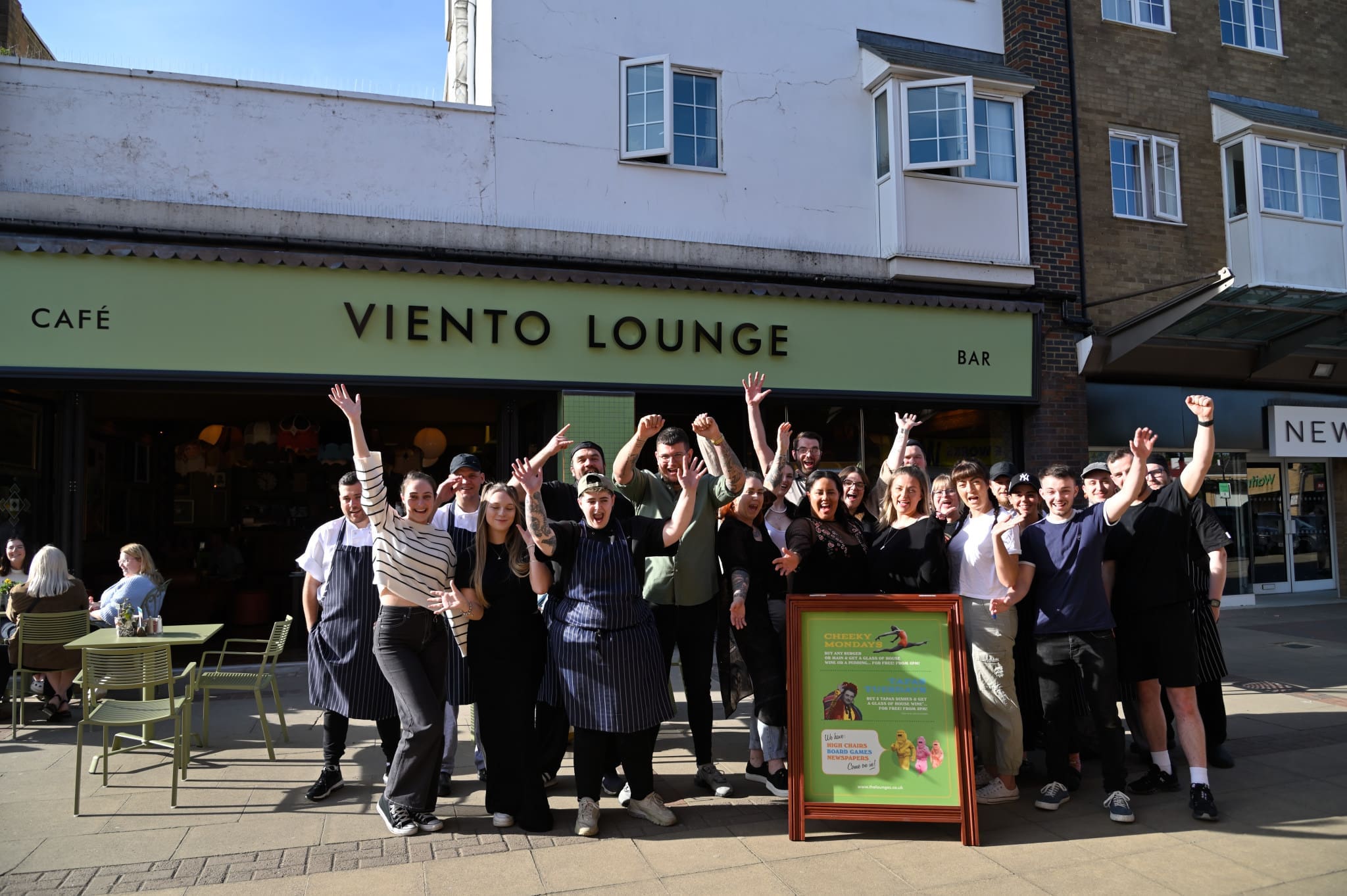 A group of people, likely staff, stand outside Viento Lounge café and bar, smiling and raising their arms in celebration in front of the vibrant green Viento Lounge storefront on a sunny day.