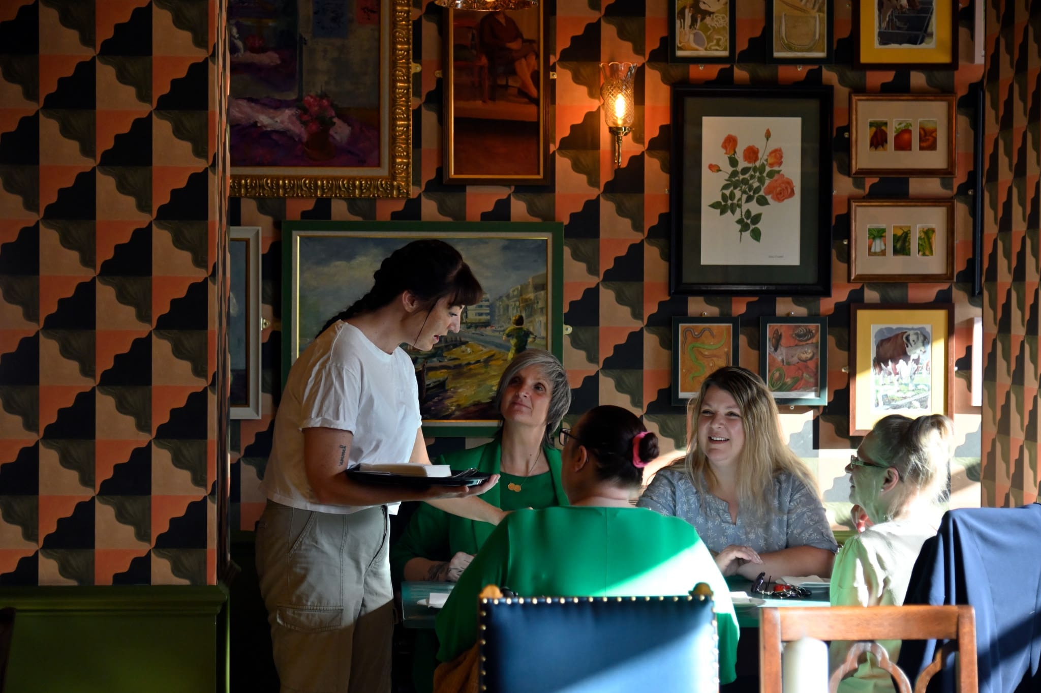 A server stands by a table taking an order from four women, one with green face paint, in the eclectic Viento Lounge. Patterned wallpaper and framed art line the walls as warm sunlight highlights the group.