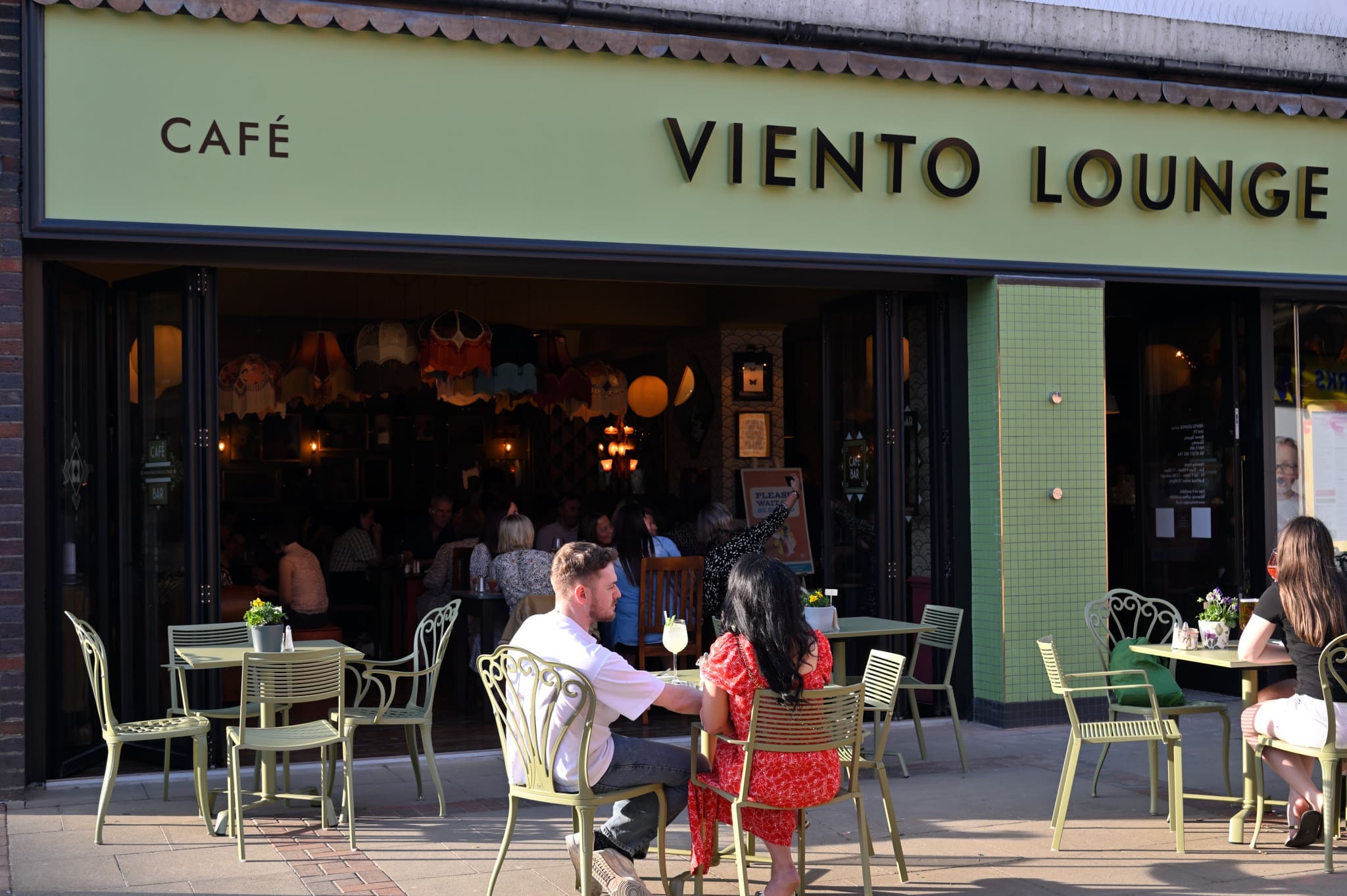 People relax at outdoor tables of Viento Lounge on a sunny day, with more guests visible inside. The café’s pale green exterior and metal pavement chairs create a welcoming spot to enjoy the vibrant atmosphere.