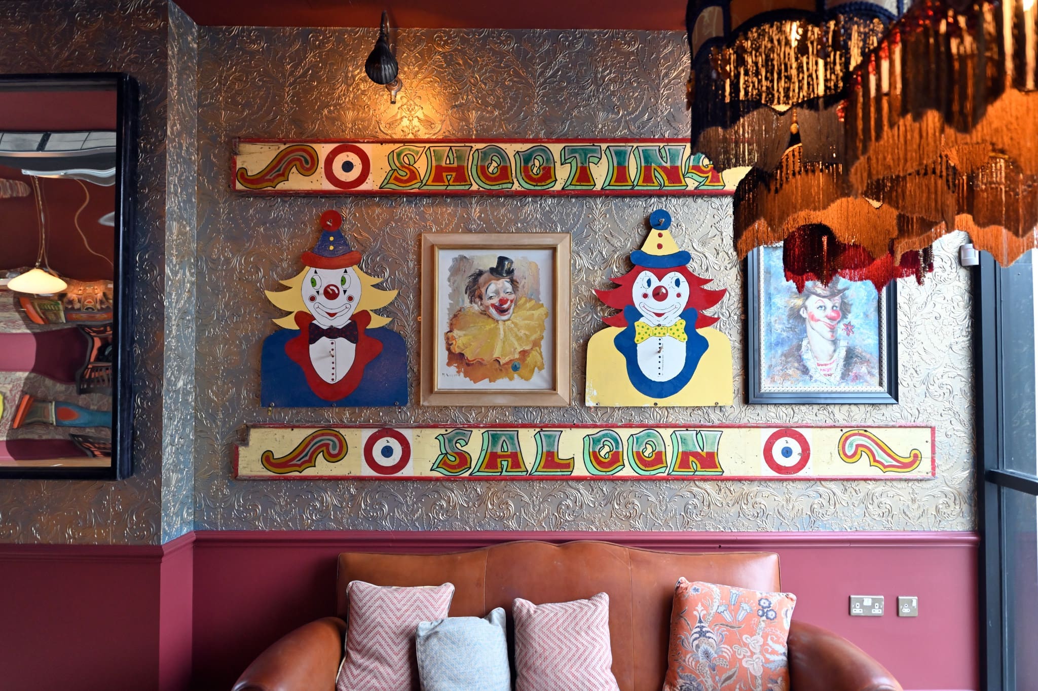 A Vintro-style wall features colorful clown artwork, a framed clown portrait, and a "Shooting Saloon" sign above a leather sofa with patterned pillows. A fringed lamp hangs in the foreground, adding to the vintage Vintro charm.