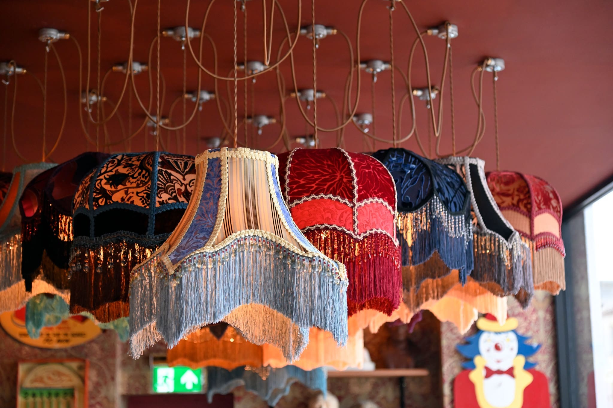 A row of vintage, Vintro fringed lampshades in various colors and patterns hangs from the ceiling by cords in a warmly lit, eclectic room. A clown painting and patterned wallpaper are visible in the background.