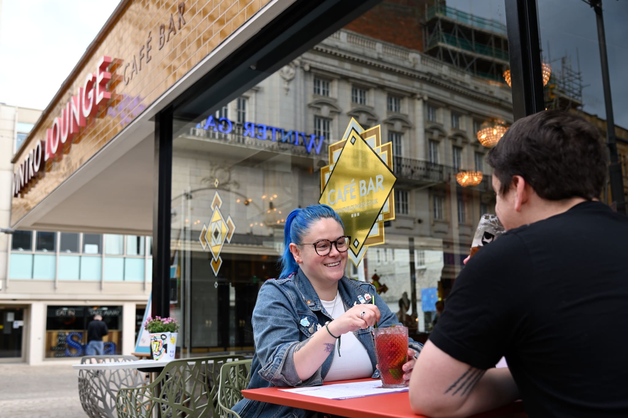 Two people sit at a Vintro café table outdoors, smiling and enjoying drinks. The woman with blue hair and glasses stirs her beverage while the café’s glass windows reflect city buildings in the background.