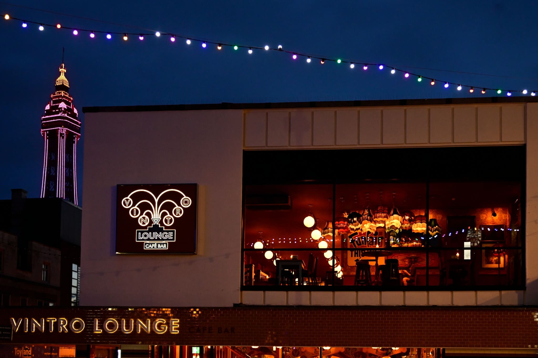 A modern Vintro lounge bar with large windows glows with warm lights at night. Colorful string lights hang above, and a tall illuminated tower stands in the background against a dark blue sky.