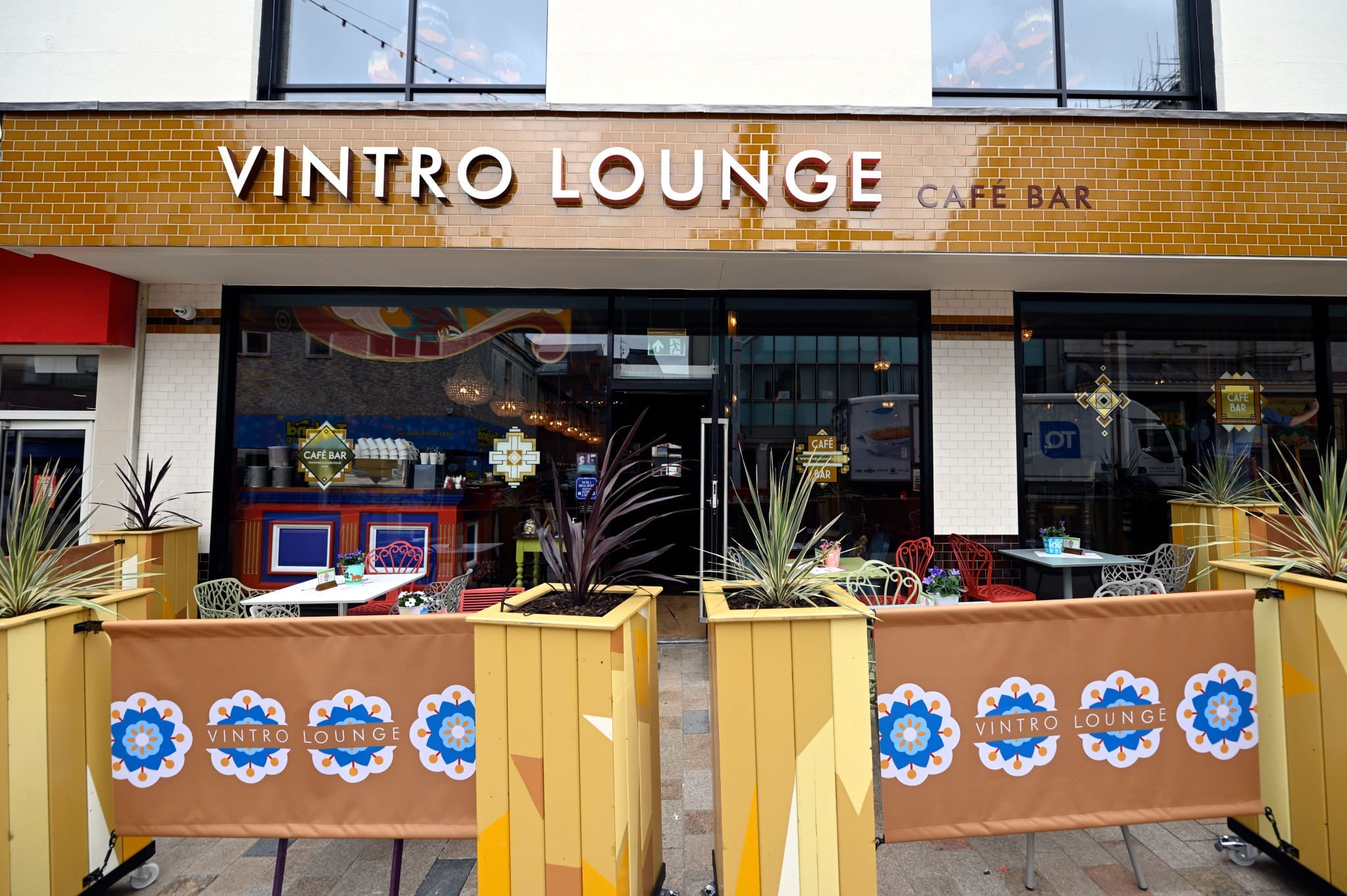 Outdoor seating area of Vintro Lounge café bar, with yellow planters, decorated barriers, and colorful chairs, in front of large glass windows displaying Vintro's stylish interior.
