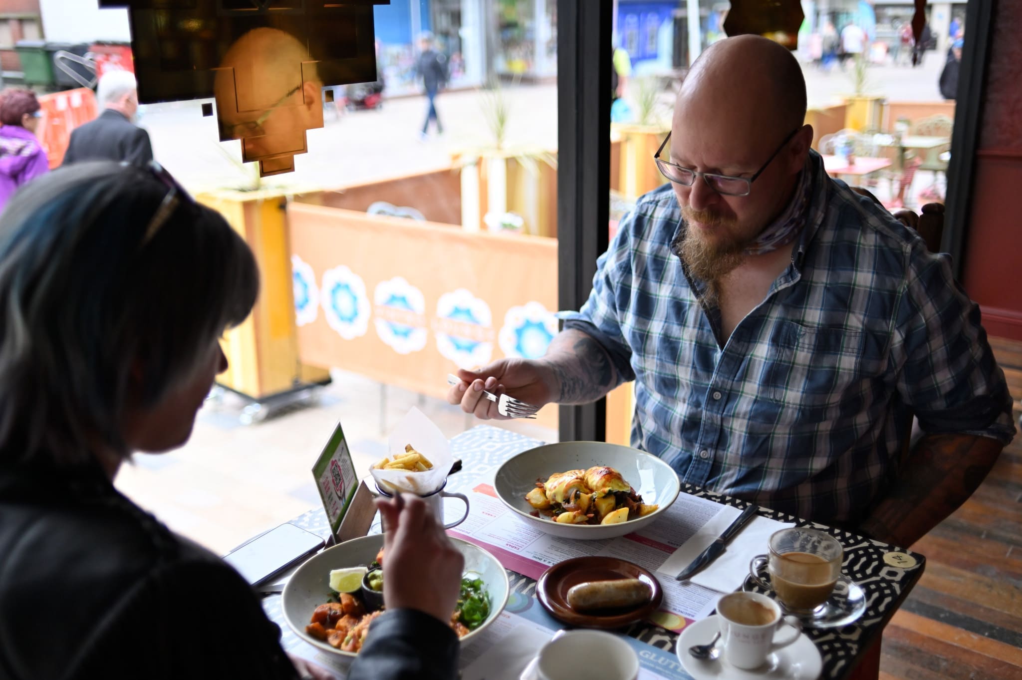 Two people sit at a table by a window in Vintro restaurant, eating meals from white bowls. One person wears a plaid shirt and glasses, and both have drinks and food in front of them. Outdoor seating is visible through the window.