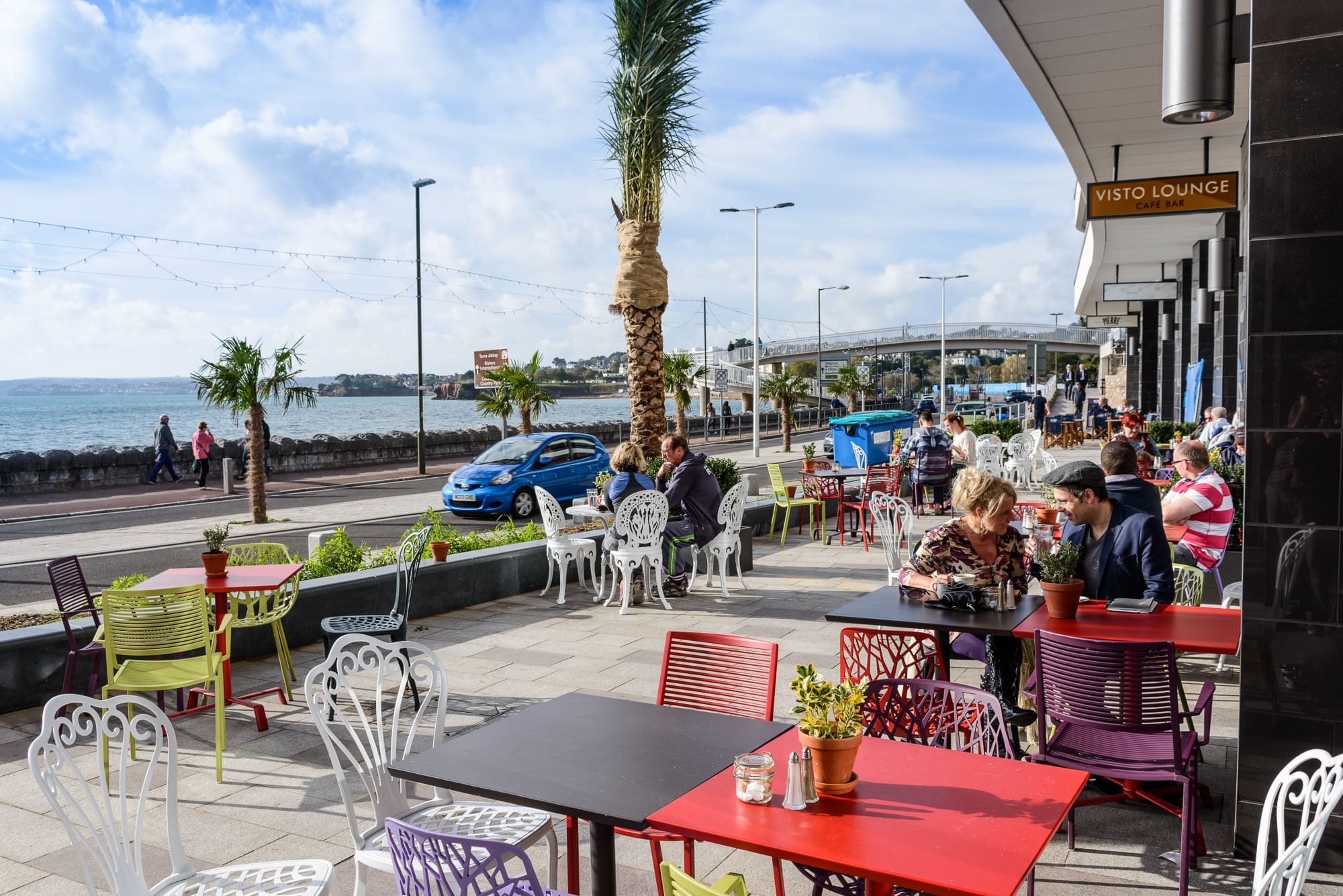 People sit at colorful outdoor tables of a seaside café on a sunny day, enjoying the Visto. A palm tree and potted plants decorate the patio, while cars and pedestrians move along the promenade in the background.