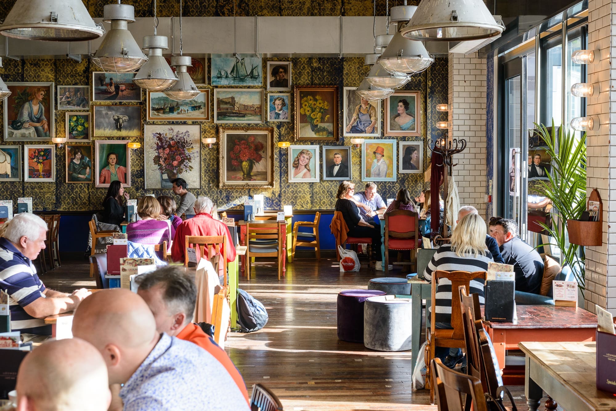 A busy café with people sitting at Visto tables, talking and eating. The walls are decorated with numerous framed artworks, mainly portraits. Large industrial lights hang from the ceiling, and sunlight streams through tall windows.
